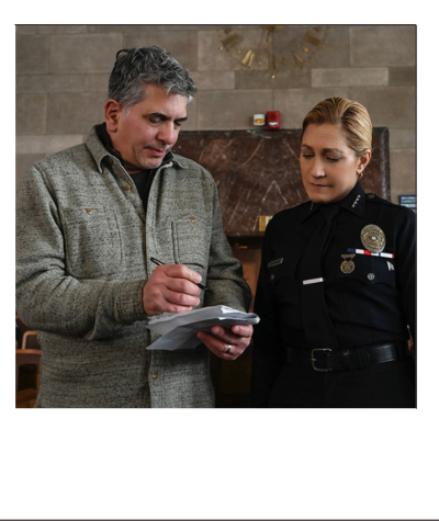 A man in a gray sweater and a woman in police uniform standing together indoors, looking at a notepad.