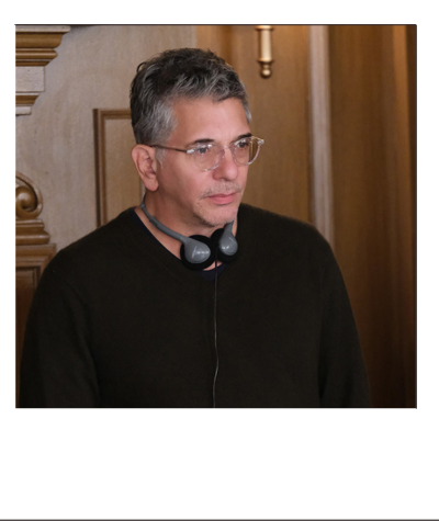A man with glasses and gray hair, wearing a black shirt, standing in front of a wooden wall with a candle holder.