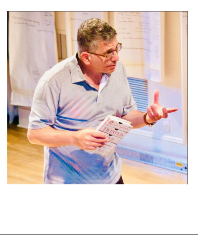A man wearing glasses and a light blue polo shirt is holding a booklet and gesturing with his right hand in a room with papers and charts on the wall.