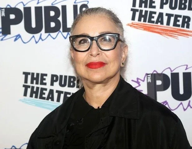 A woman with gray hair, wearing glasses and red lipstick, posing in front of a backdrop with the words 'THE PUBLIC THEATER' and 'THE PUBLIC' in various styles and colors.