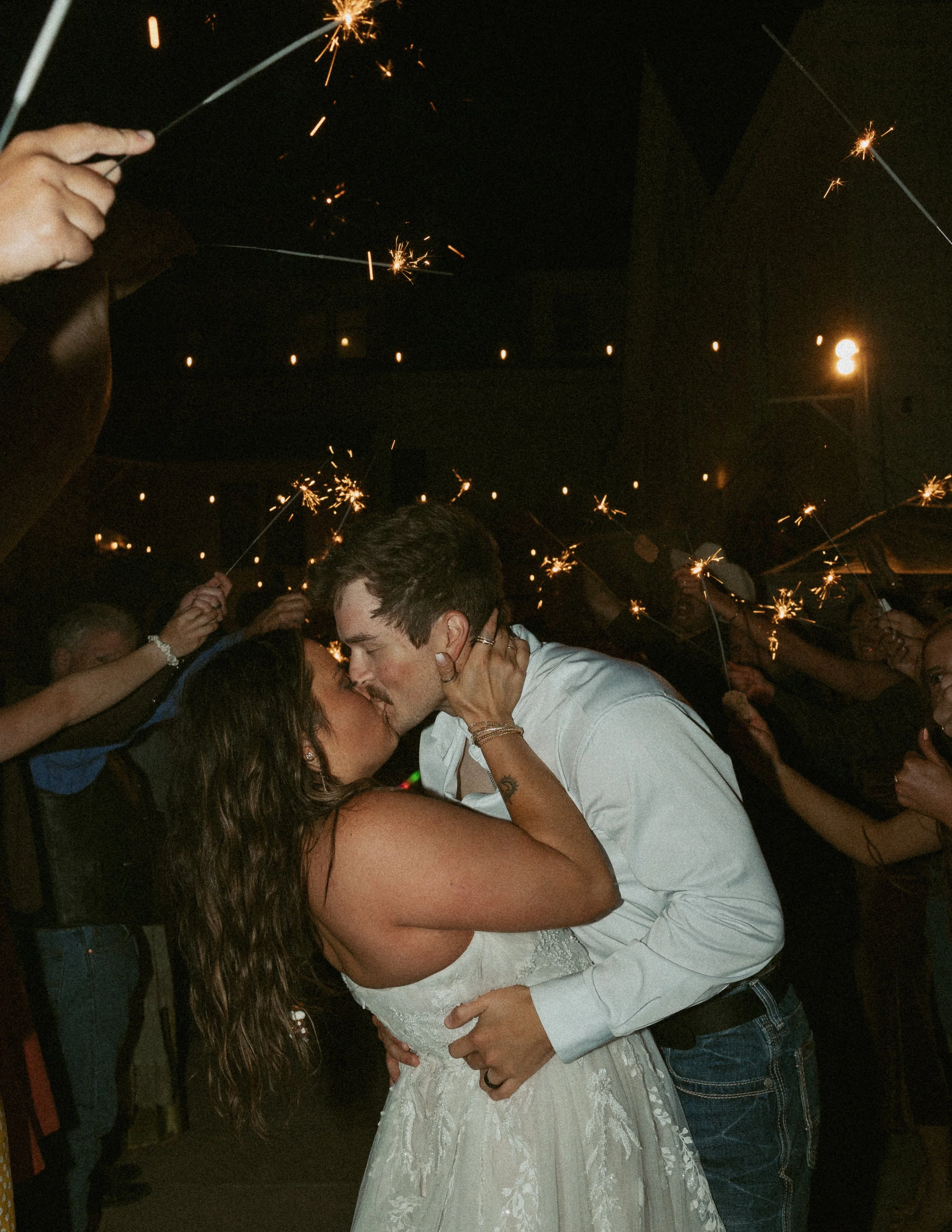 A couple sharing a kiss at a wedding reception, surrounded by people holding sparklers and celebrating in the dark.