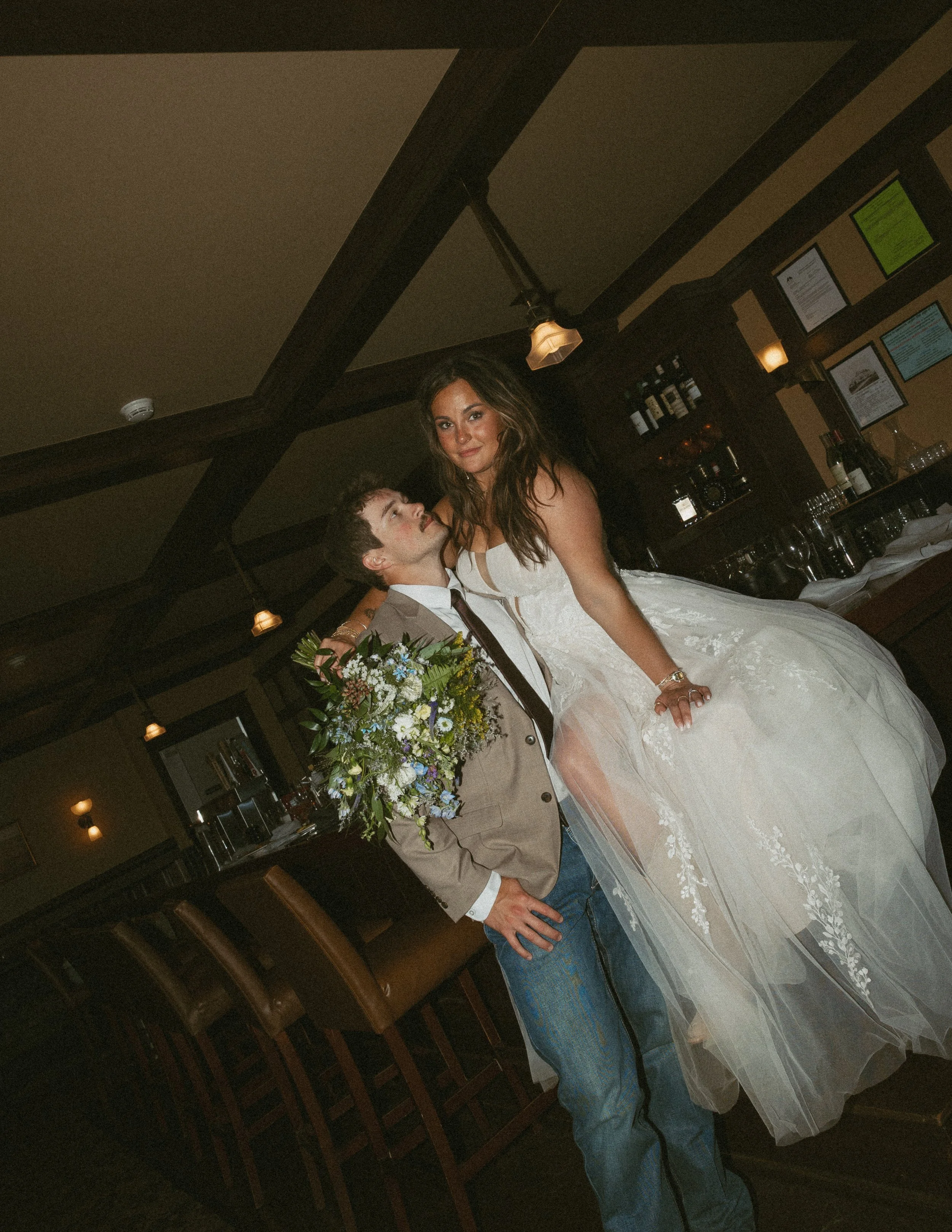 A bride in a white wedding gown and a groom in a beige jacket and blue jeans stand together in a dimly lit room with wood-paneled walls and framed certificates. The bride is holding a bouquet of flowers, and the couple is smiling at each other.