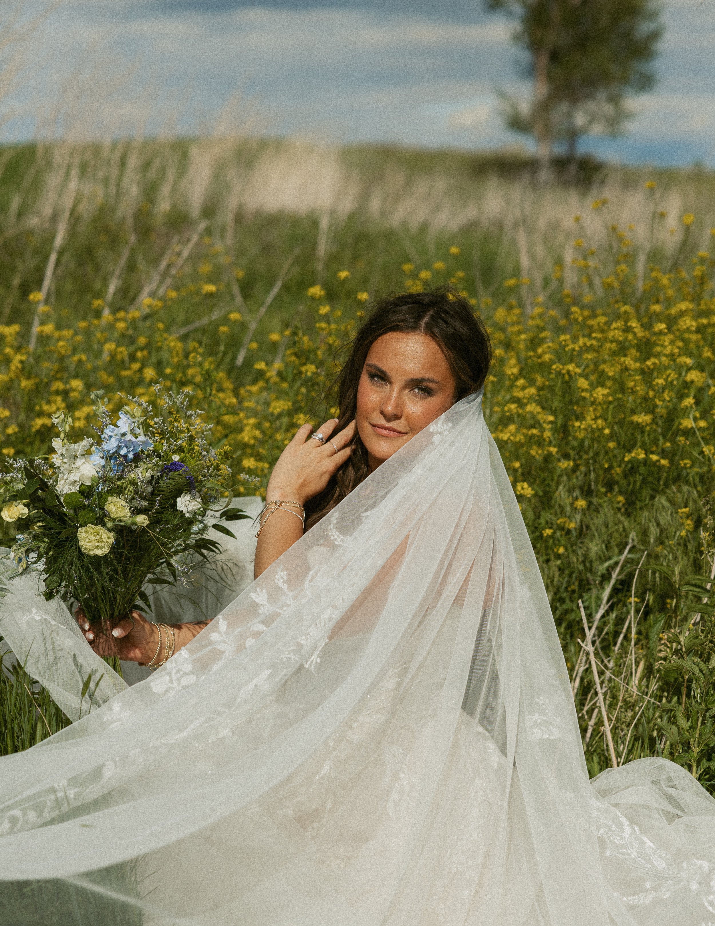 A woman with dark brown hair and light skin in a white dress holding a bouquet of flowers, standing in a field of yellow and white flowers with a tree and blue sky in the background.
