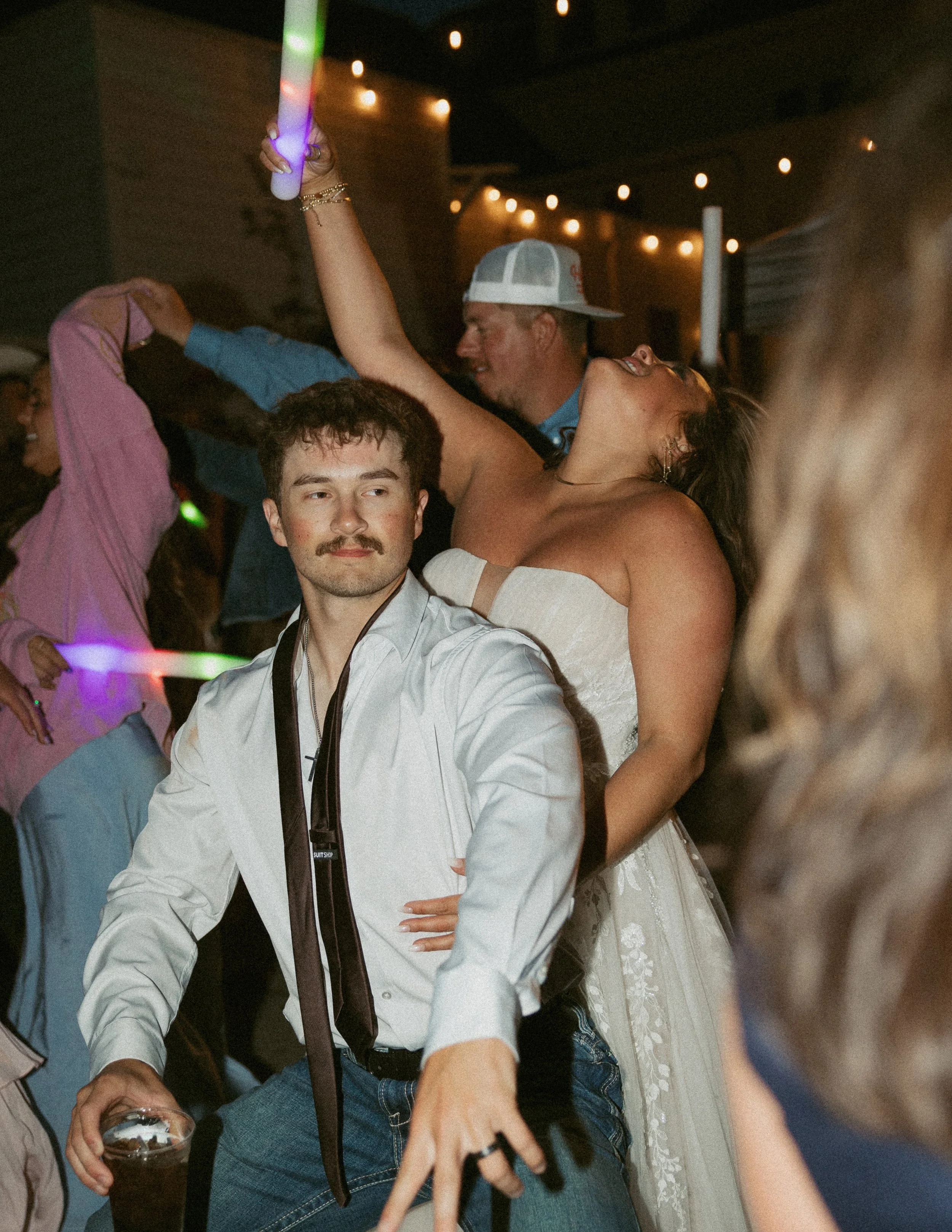 People dancing and socializing at a party or celebration. Prominent person in the foreground with a mustache wearing a white shirt and black tie, with a camera strap around his neck. A woman in a white dress and woman in a cap are also visible, with 
