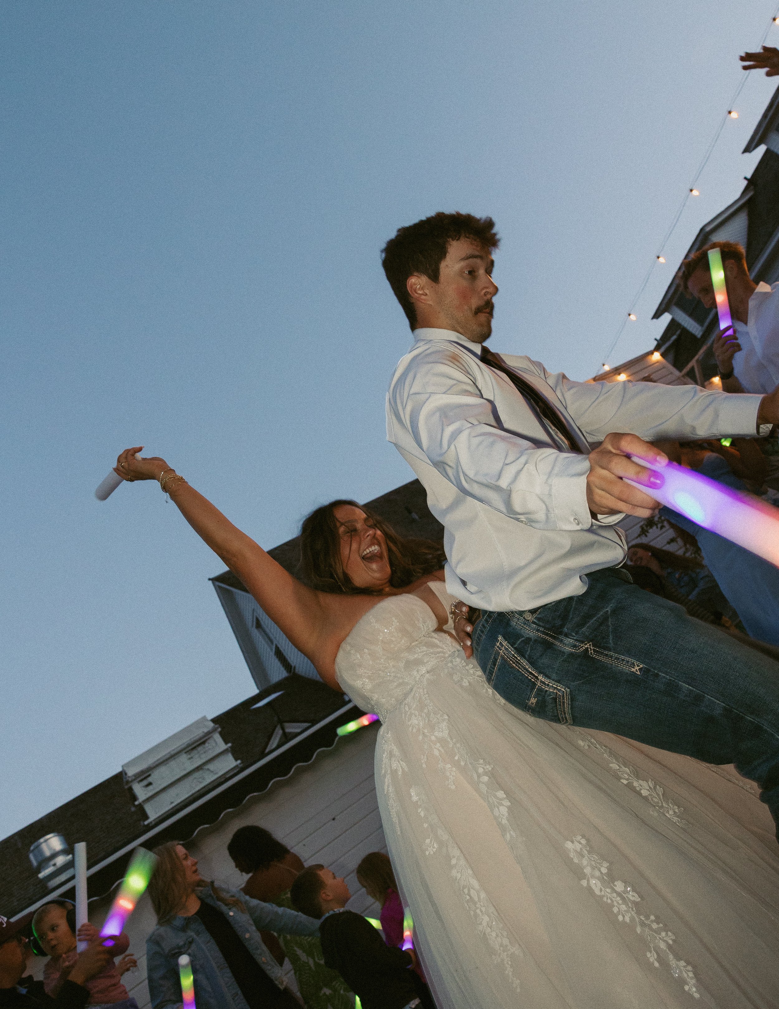 A bride in a white wedding dress dancing at an outdoor wedding reception during twilight, with guests holding glow sticks nearby.