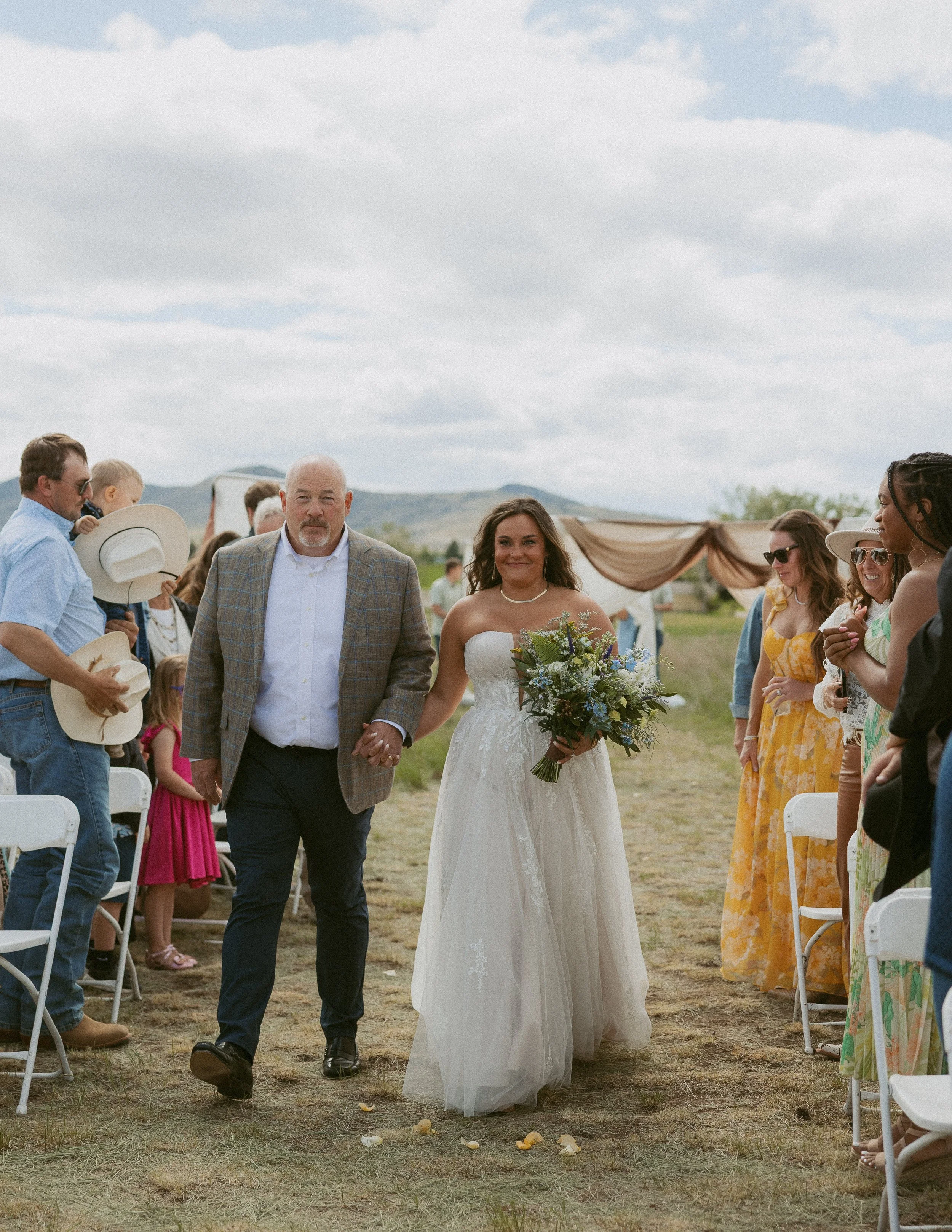 A bride holding a bouquet of flowers walks down the aisle at an outdoor wedding ceremony, accompanied by an older man, with guests seated on both sides.