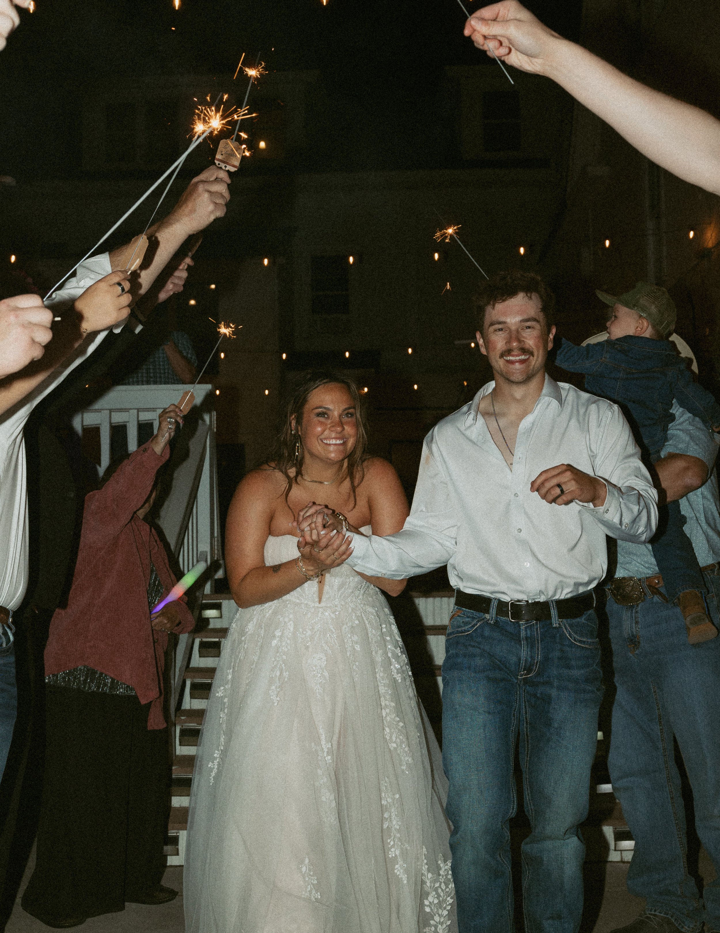 People holding sparklers at a celebration, with a smiling couple in the center, a woman in a white dress and a man in a white shirt.