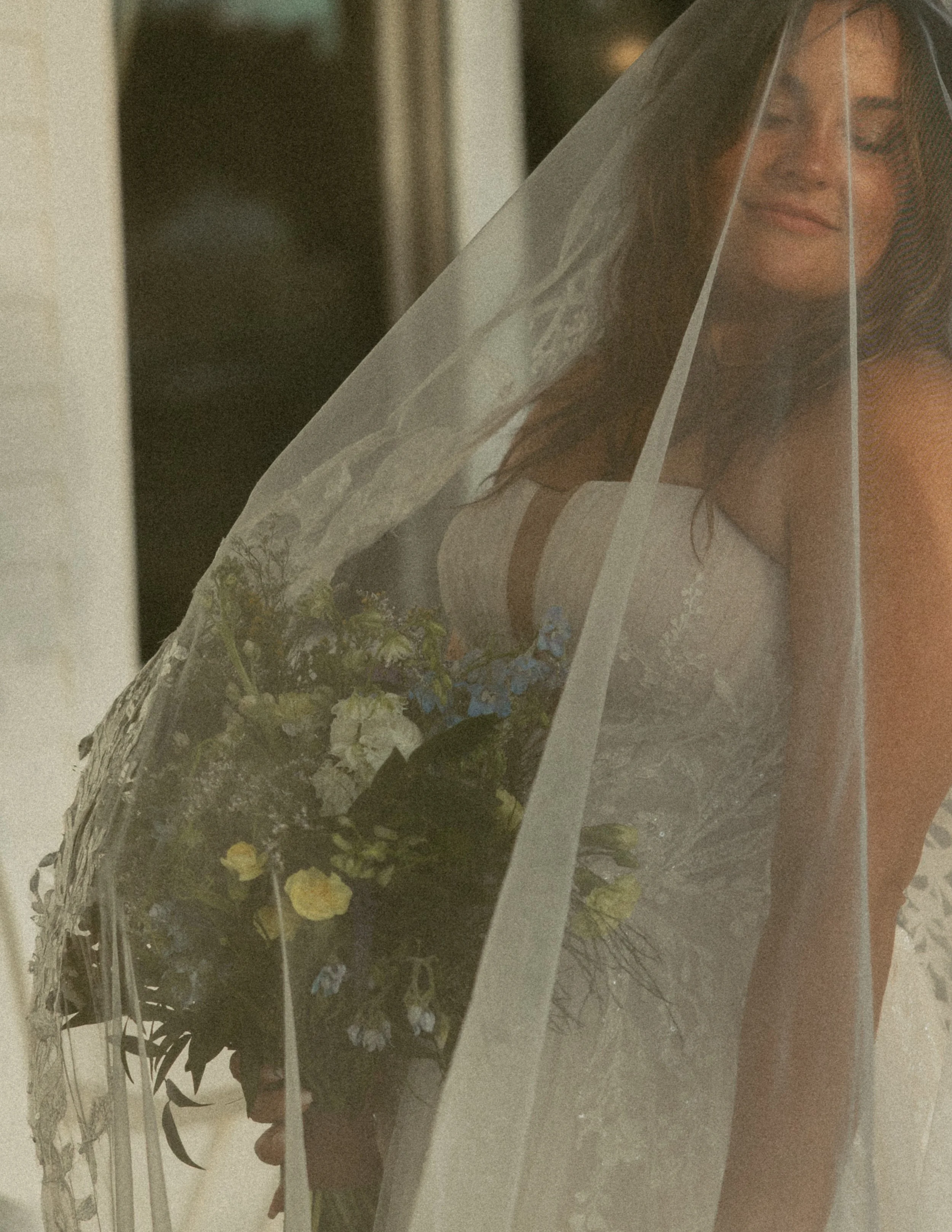 A woman with long brown hair, wearing a white top, is looking through a glass window or door with a sheer curtain or veil partially covering her face, smiling softly.