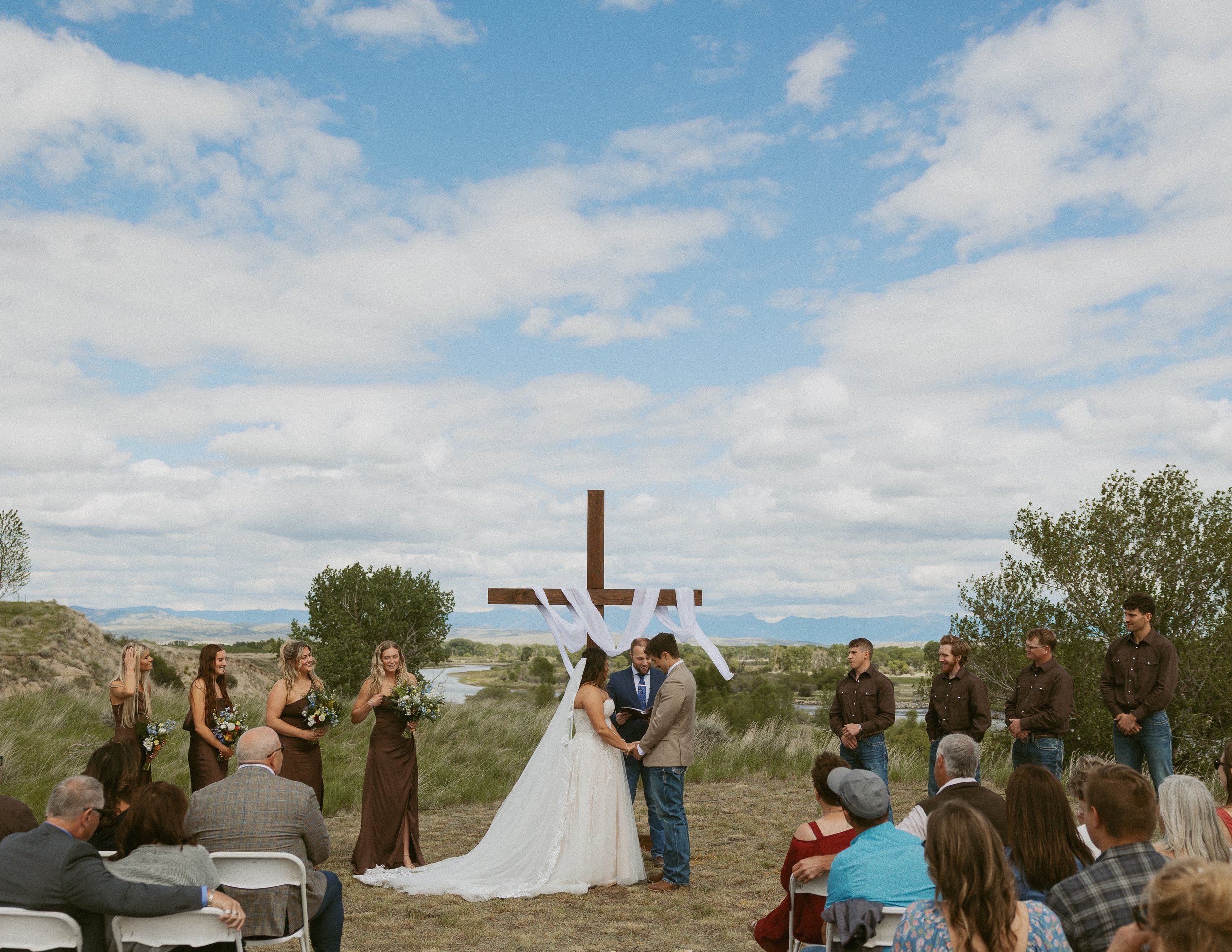 Outdoor wedding ceremony with a cross decorated with white ribbons as the backdrop, set in a grassy area under a cloudy sky, with guests seated and standing around and a wedding couple exchanging vows.