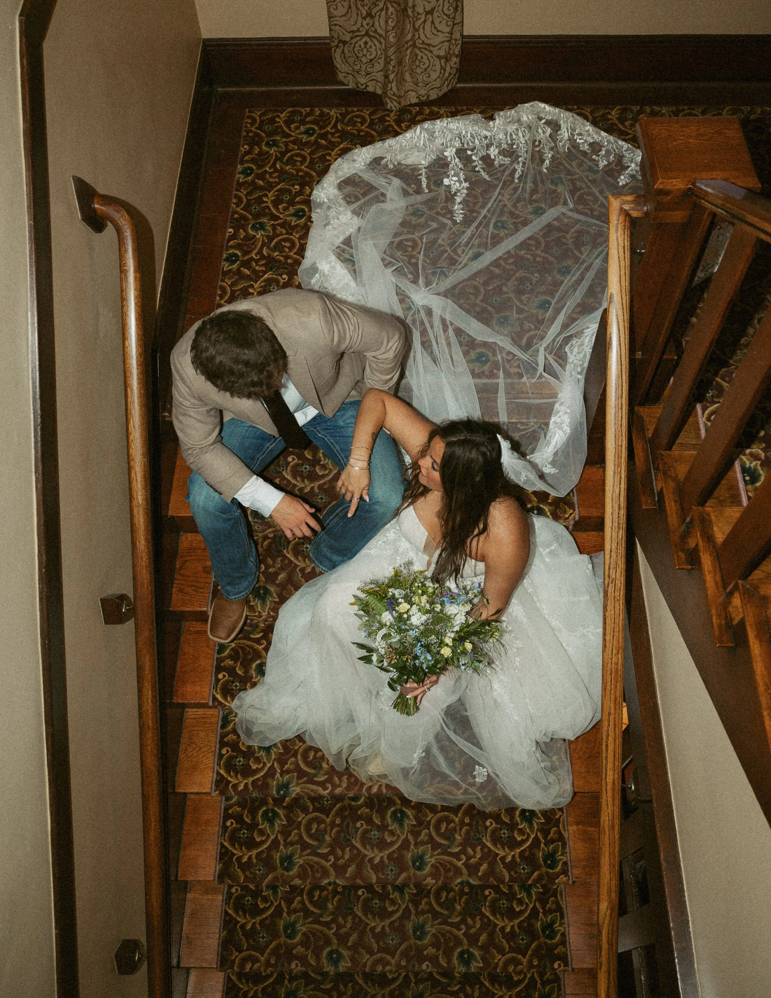 A bride in a white wedding dress holding a bouquet, lying on a wedding aisle runner with her eyes closed, while a groom in a beige blazer and jeans kneels beside her, touching her hand, in a church with wooden pews and an ornate patterned carpet.