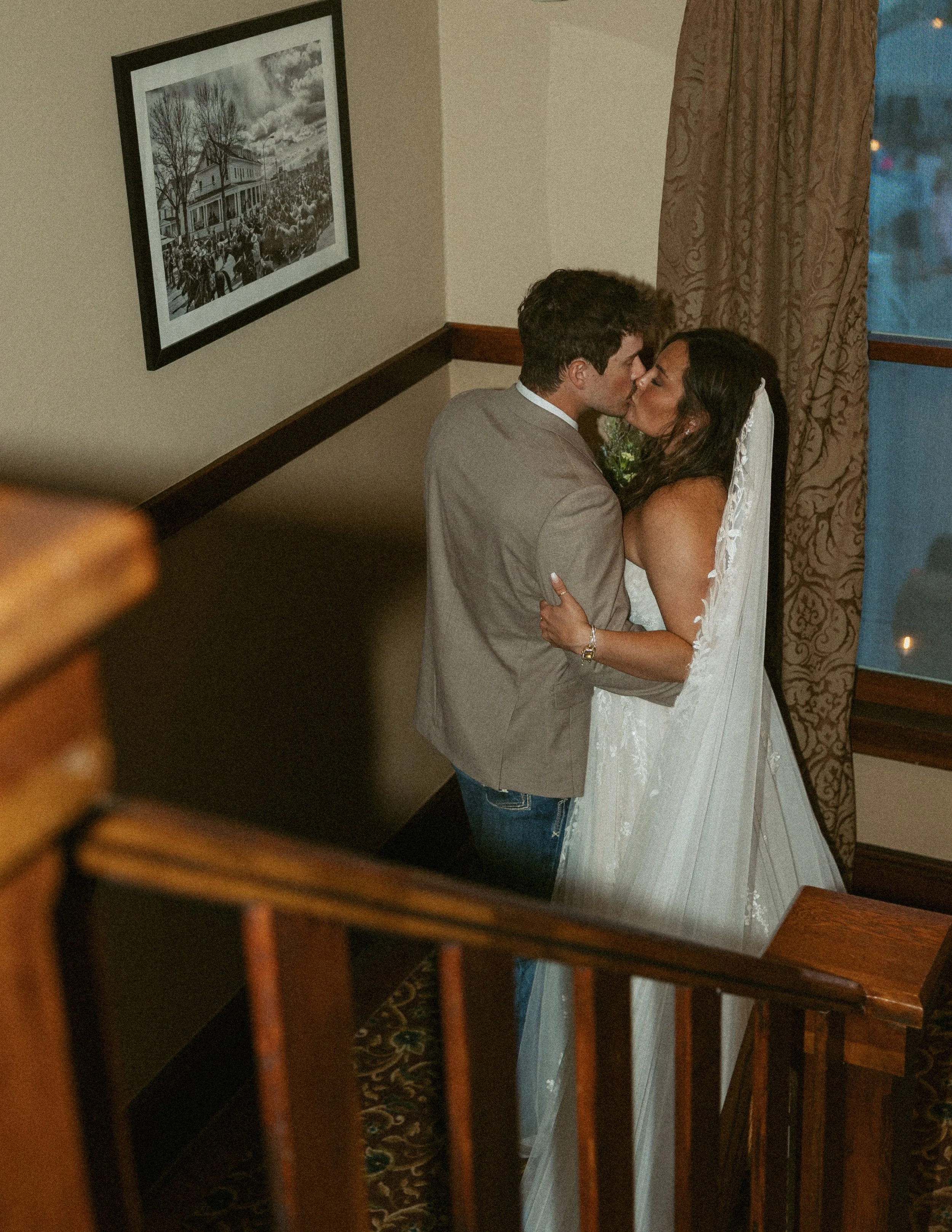 A person holding a smartphone and taking a photo of a couple kissing in a mirror. The couple is dressed in wedding attire, with the bride in a white gown and veil and the groom in a light gray suit. A framed black-and-white photograph of a historic s