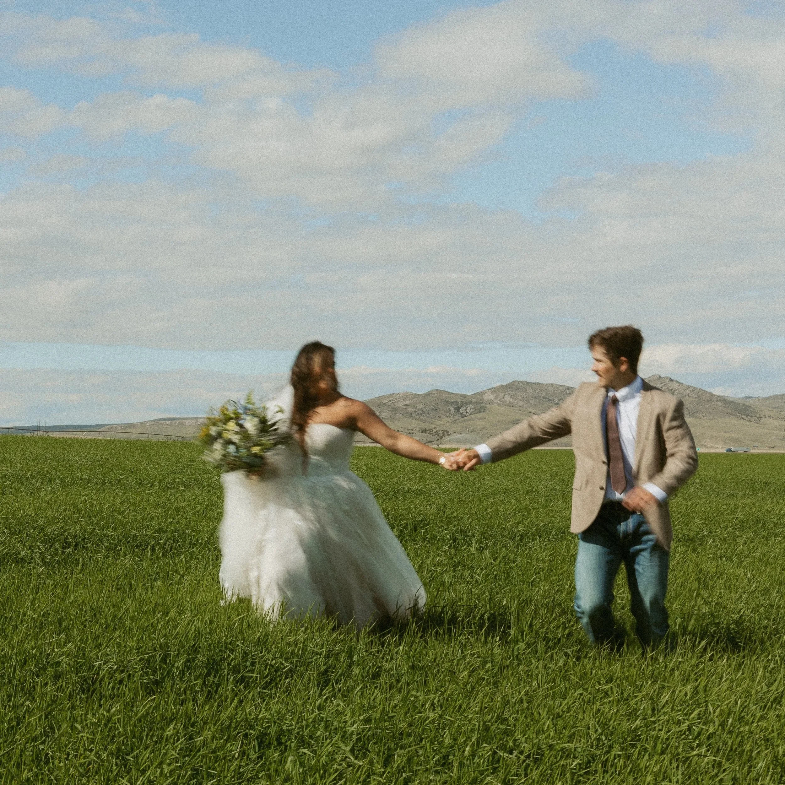 A man in a tan blazer and a woman in a white wedding dress holding hands and walking in a green field with mountains in the background, under a partly cloudy sky.