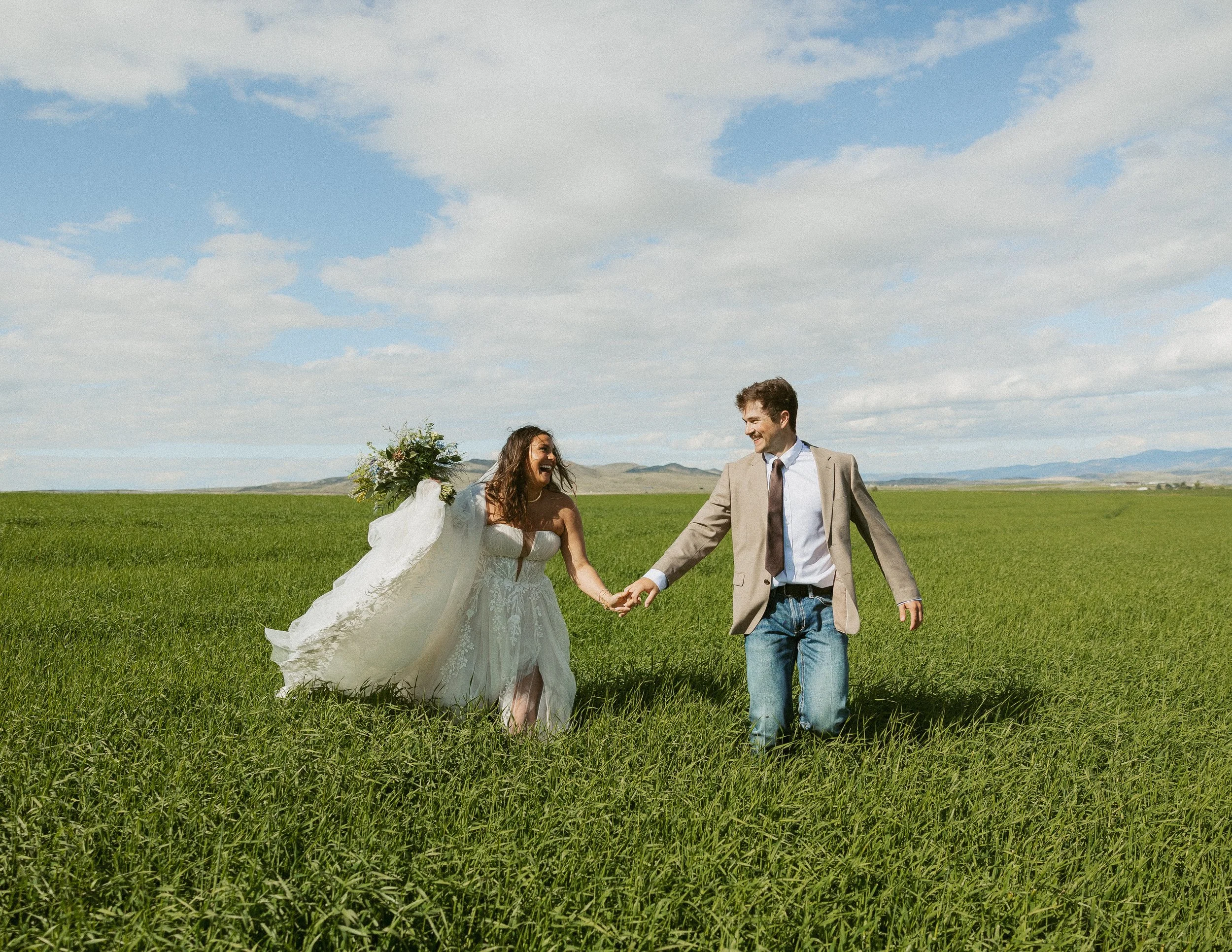 A bride and groom holding hands and smiling in a green field under a partly cloudy sky, with distant hills in the background.