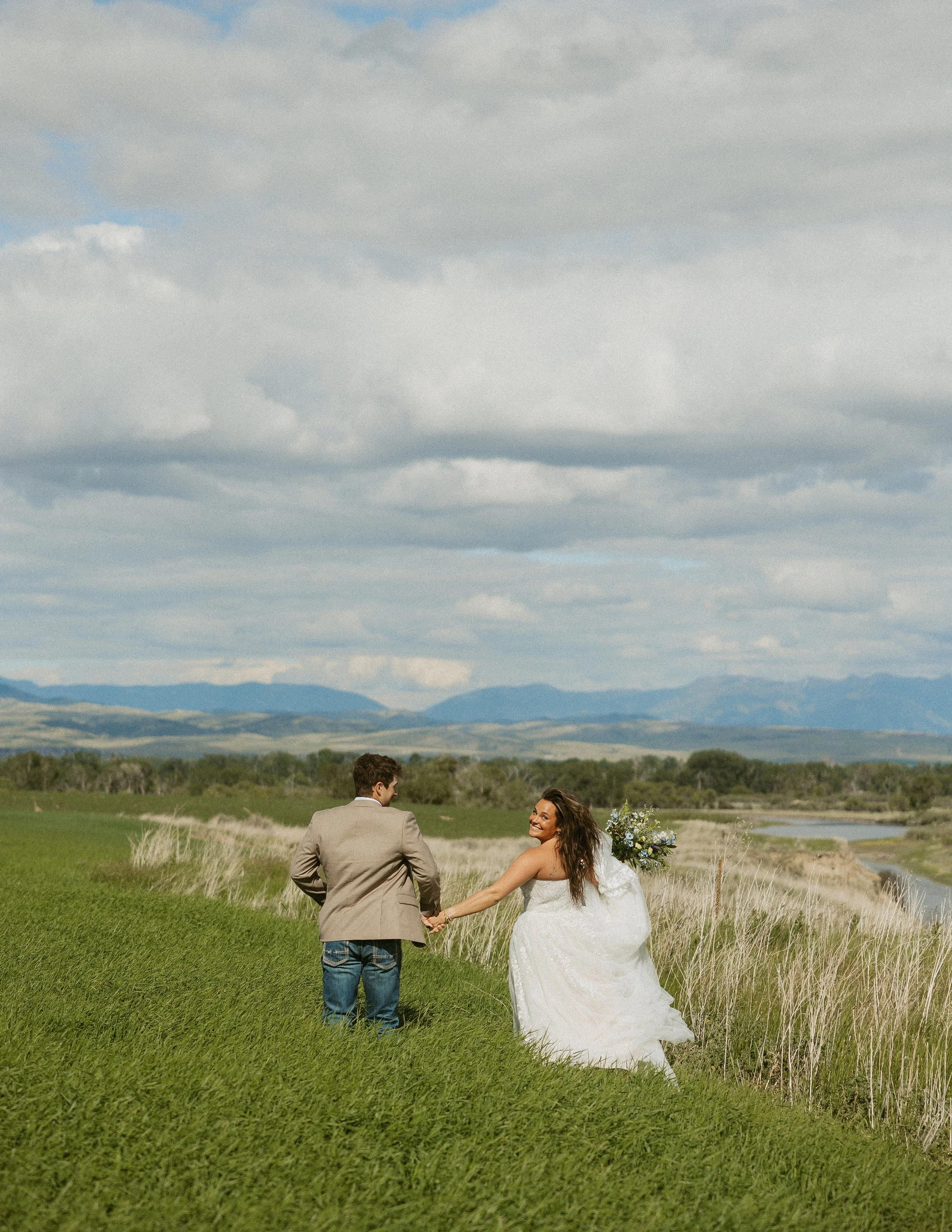 A bride and groom holding hands in a grassy field outdoors, with mountains and a cloudy sky in the background, during a wedding ceremony.