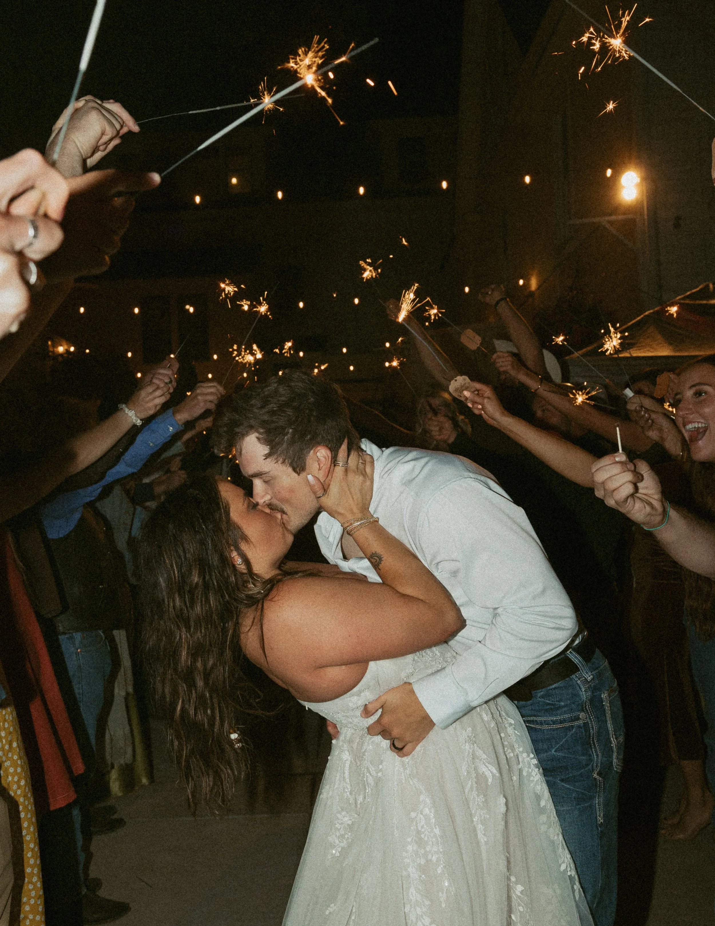 A couple kissing at a celebration, surrounded by people holding sparklers at night.