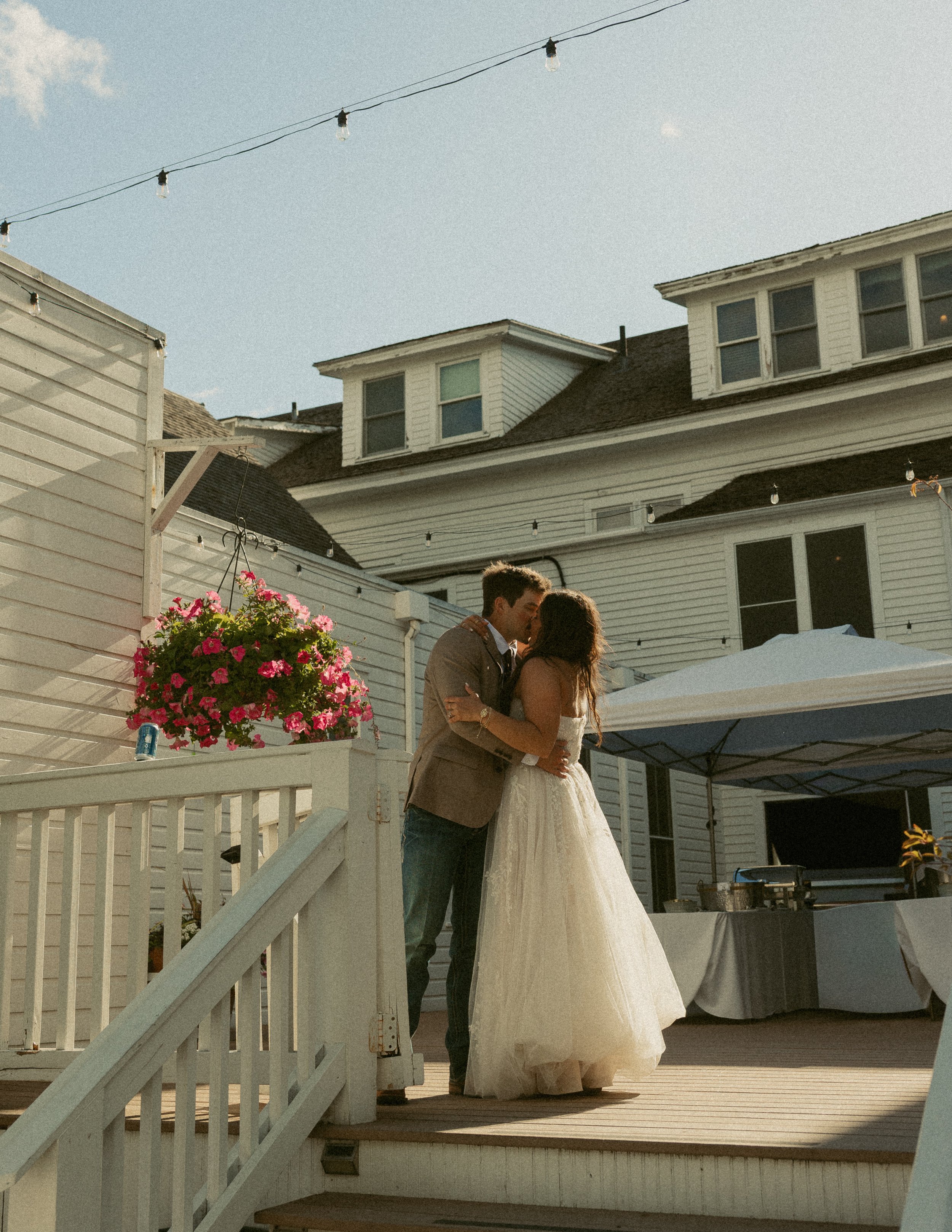 A bride and groom embrace for a kiss on a wooden deck at an outdoor wedding reception during daytime, with string lights overhead, a white house in the background, and a large white umbrella outside.