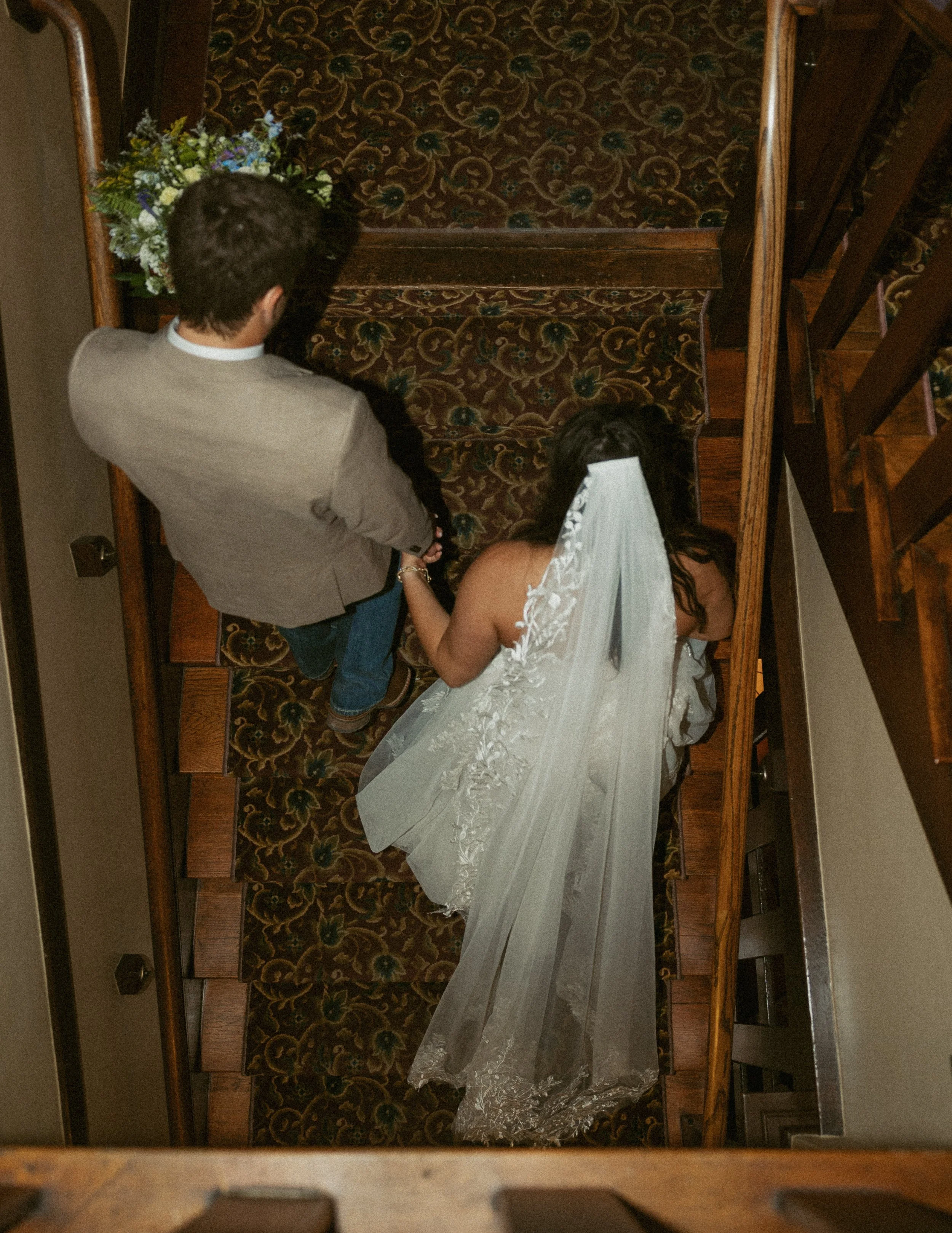 A man and woman lying on the stairs, the woman is wearing a white dress with lace details and the man is wearing a gray jacket. There is a bouquet of flowers placed near the man.
