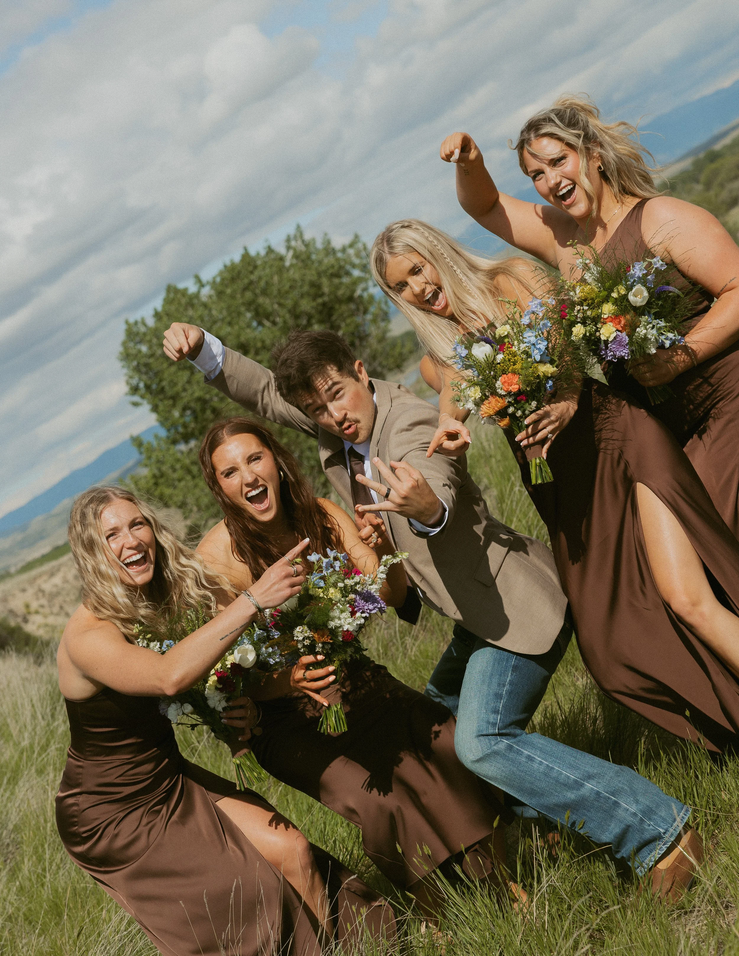 A group of five smiling people posing outdoors in a field, holding colorful bouquets of flowers, with a backdrop of mountains and trees under a partly cloudy sky. They are making playful gestures and appear joyful.