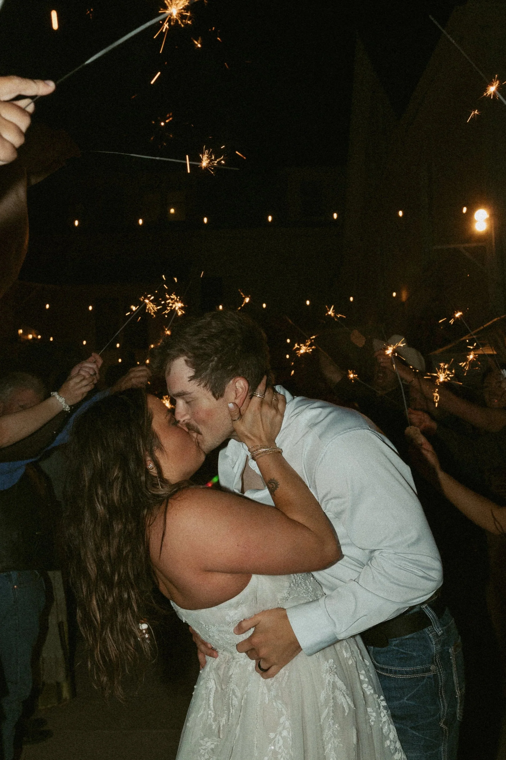 A couple is kissing at a celebration, surrounded by people holding sparklers in a dark setting. The woman is wearing a white wedding dress and the man is in a white shirt.