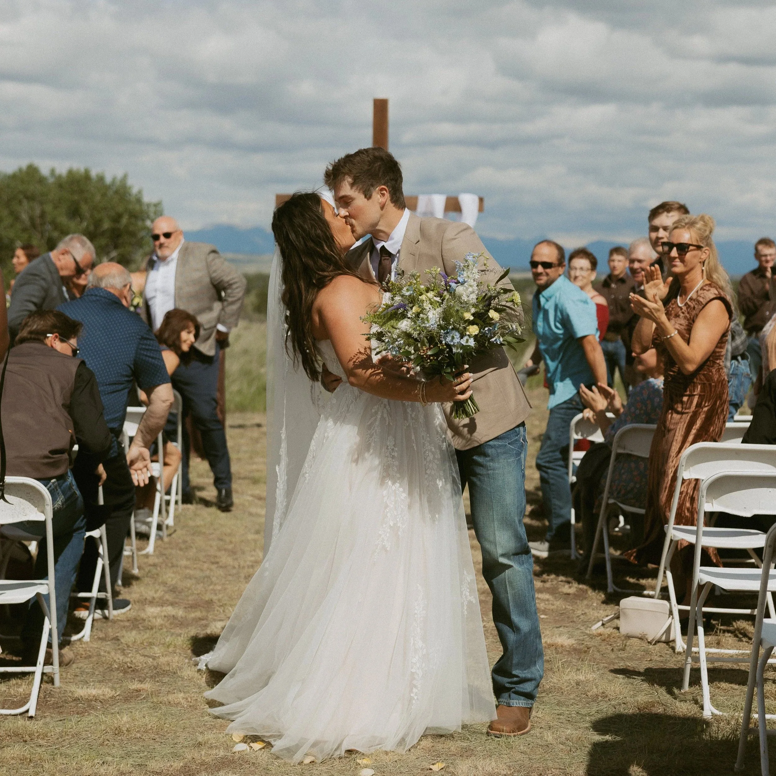 A wedding ceremony outdoors with a couple sharing a kiss, surrounded by seated and standing guests, with a large wooden cross in the background and cloudy skies.