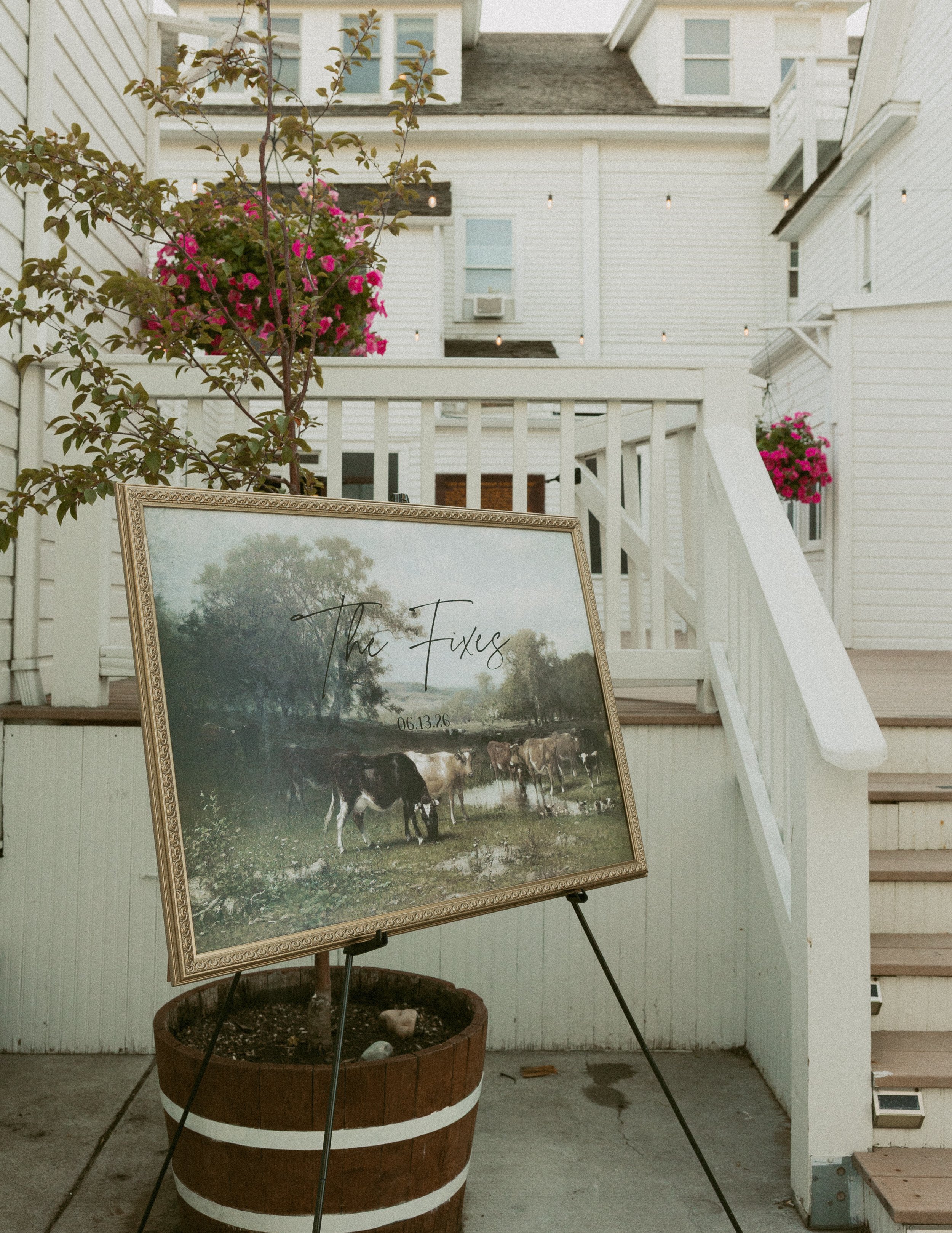 A framed photo on a stand shows a landscape with trees and a group of cows, propped up against a large potted plant with pink flowers, on a porch with white wooden stairs, railing, and house exterior.
