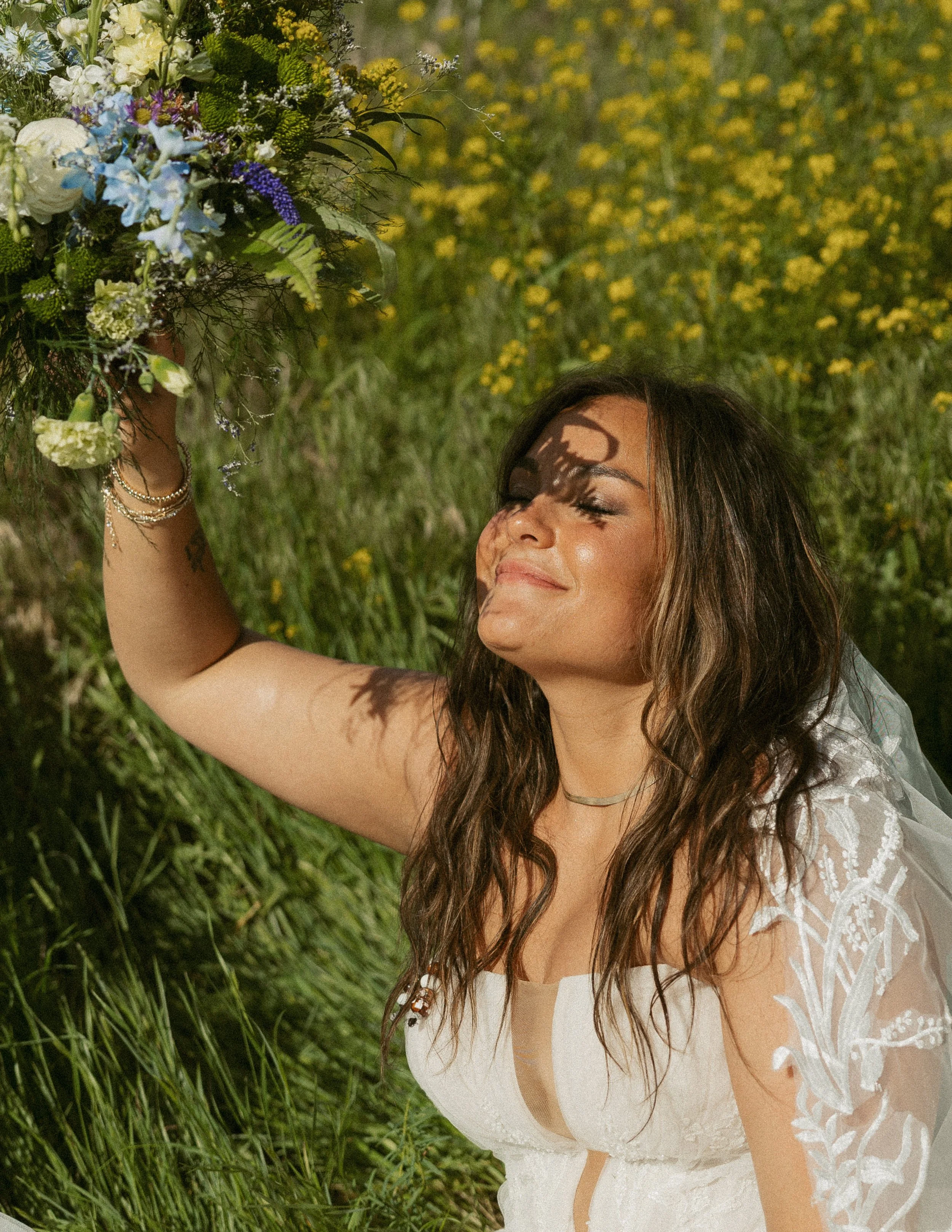 A woman with long, wavy dark hair and light brown skin is outdoors in a grassy area with yellow flowers in the background. She is smiling with her eyes closed, wearing a white lace dress and jewelry, and holding a bouquet of flowers.
