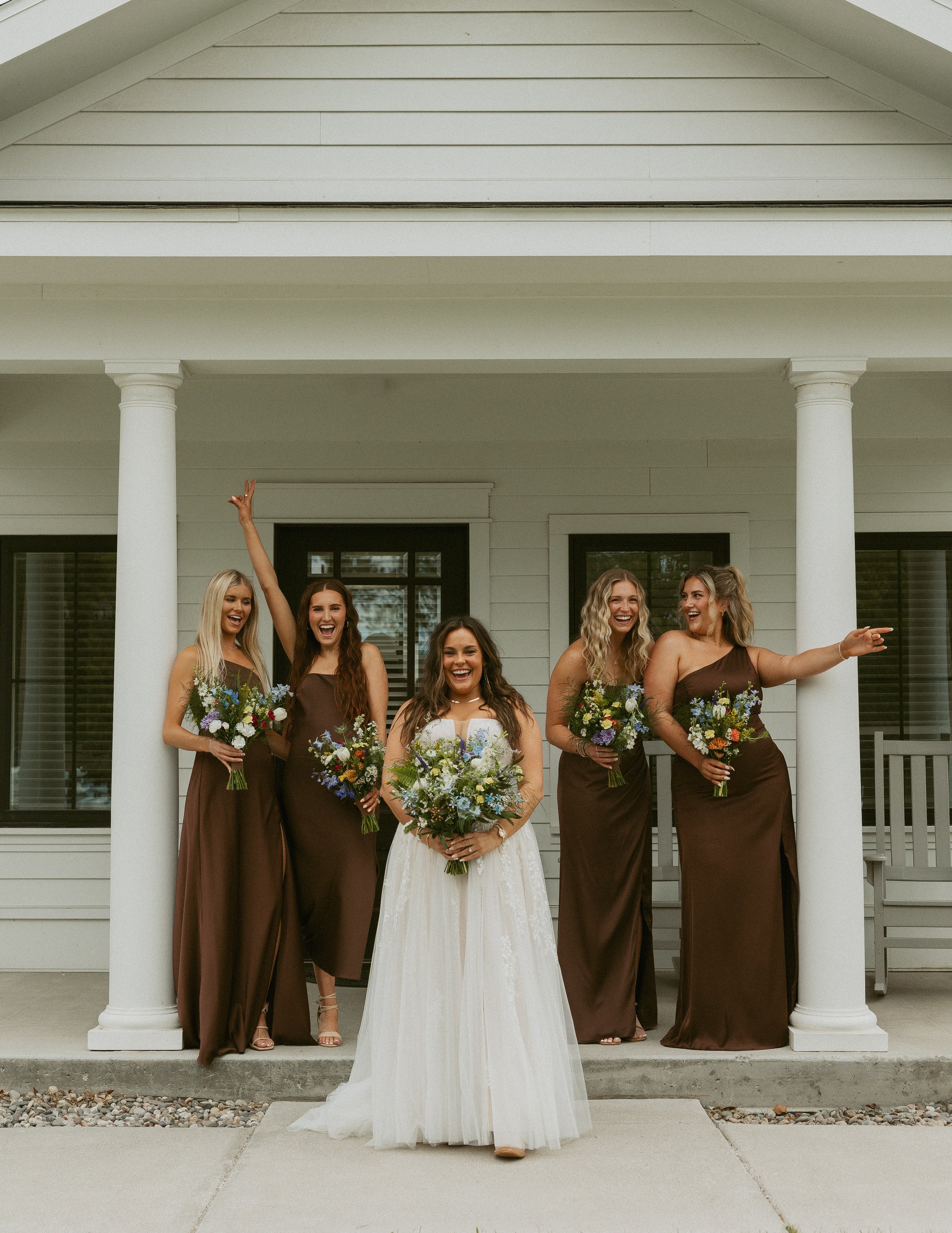 A bride in a white wedding dress standing in front of her bridesmaids on a porch with columns. The bridesmaids are wearing brown dresses and holding bouquets of flowers, and they are smiling and laughing.