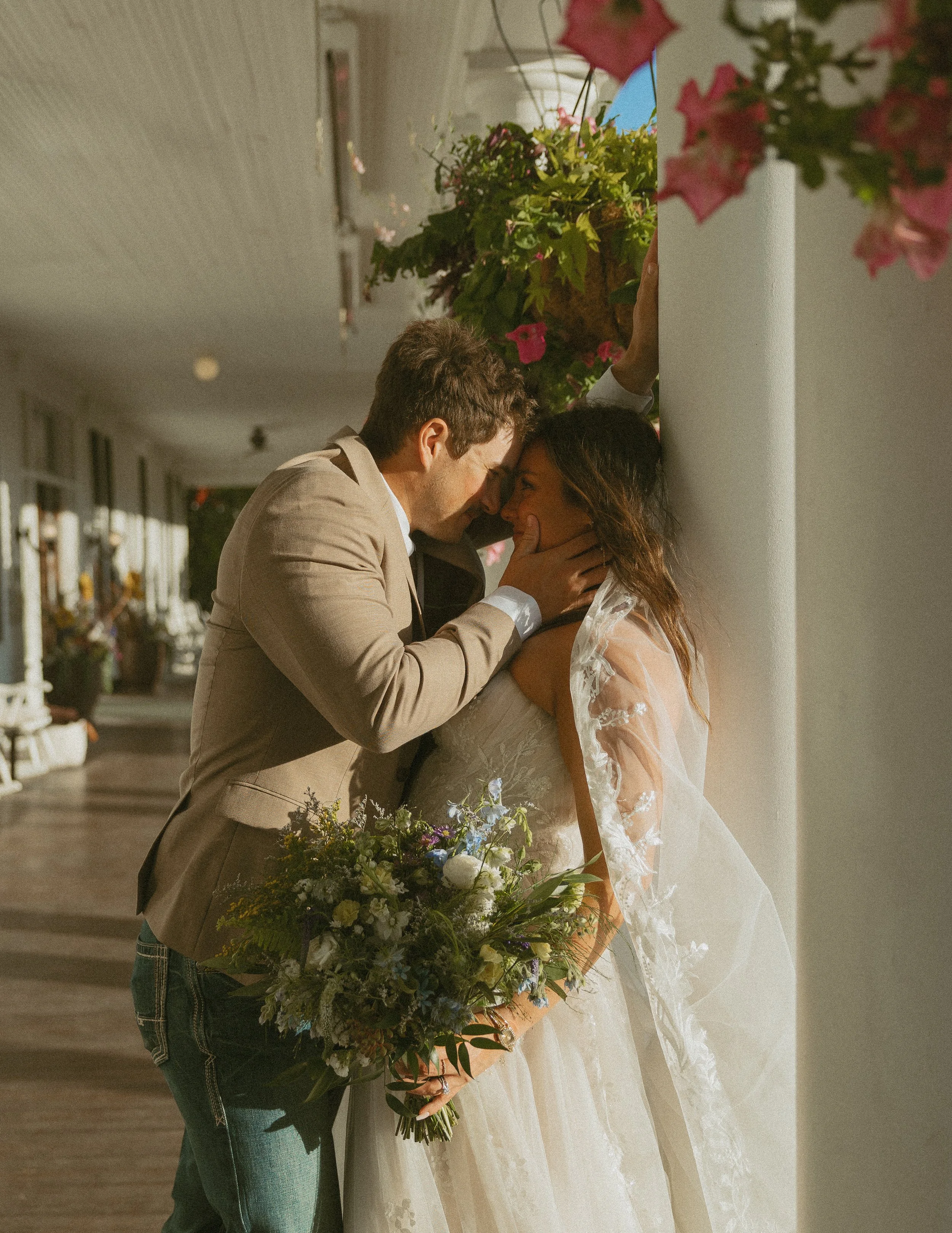 A man and woman at a wedding, with the man holding a large bouquet of flowers, leaning in close to the woman who is smiling, both touching noses, near a white pillar with pink flowers hanging above.