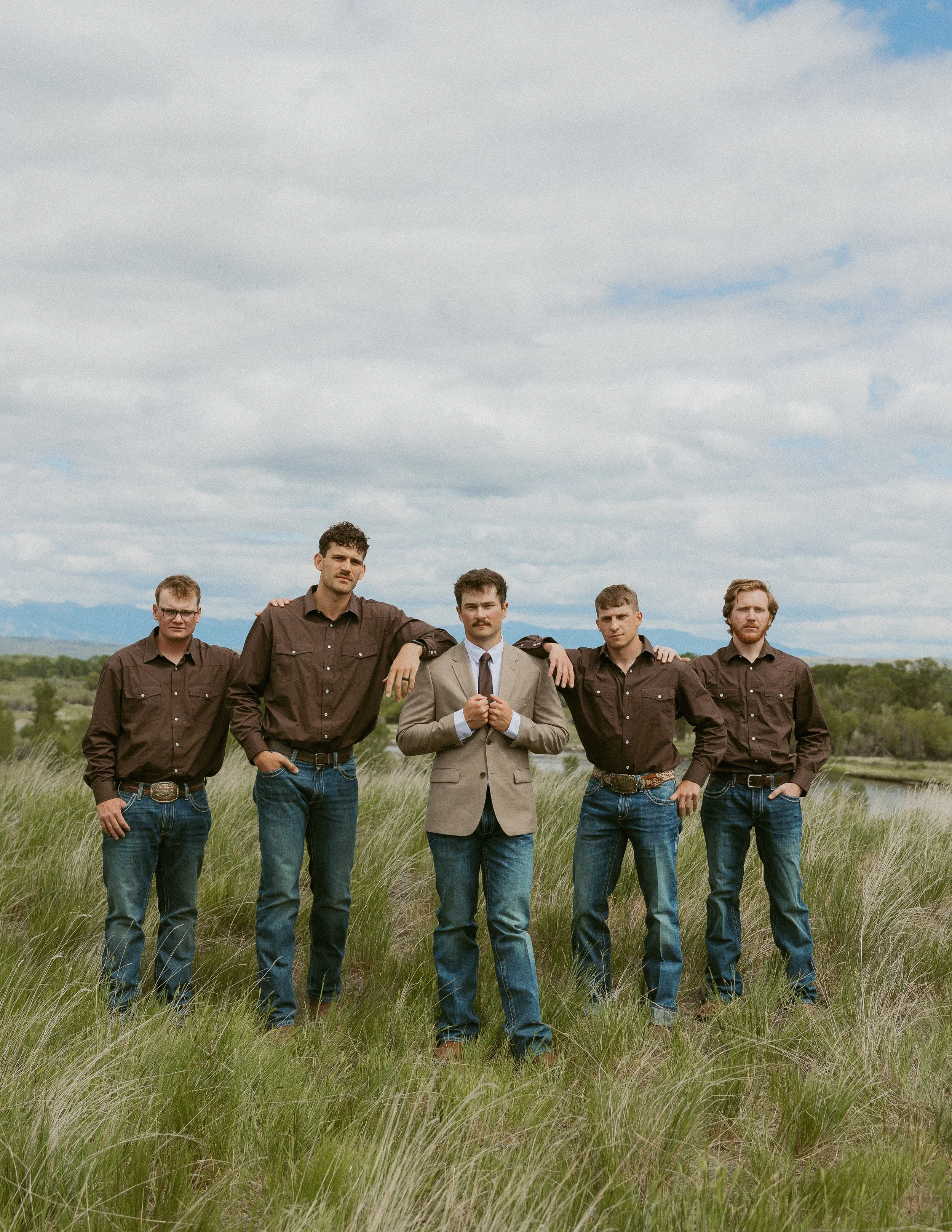 Group of six men standing in a grassy field under a cloudy sky, dressed in casual and semi-formal attire, with one man in a beige suit in the center.