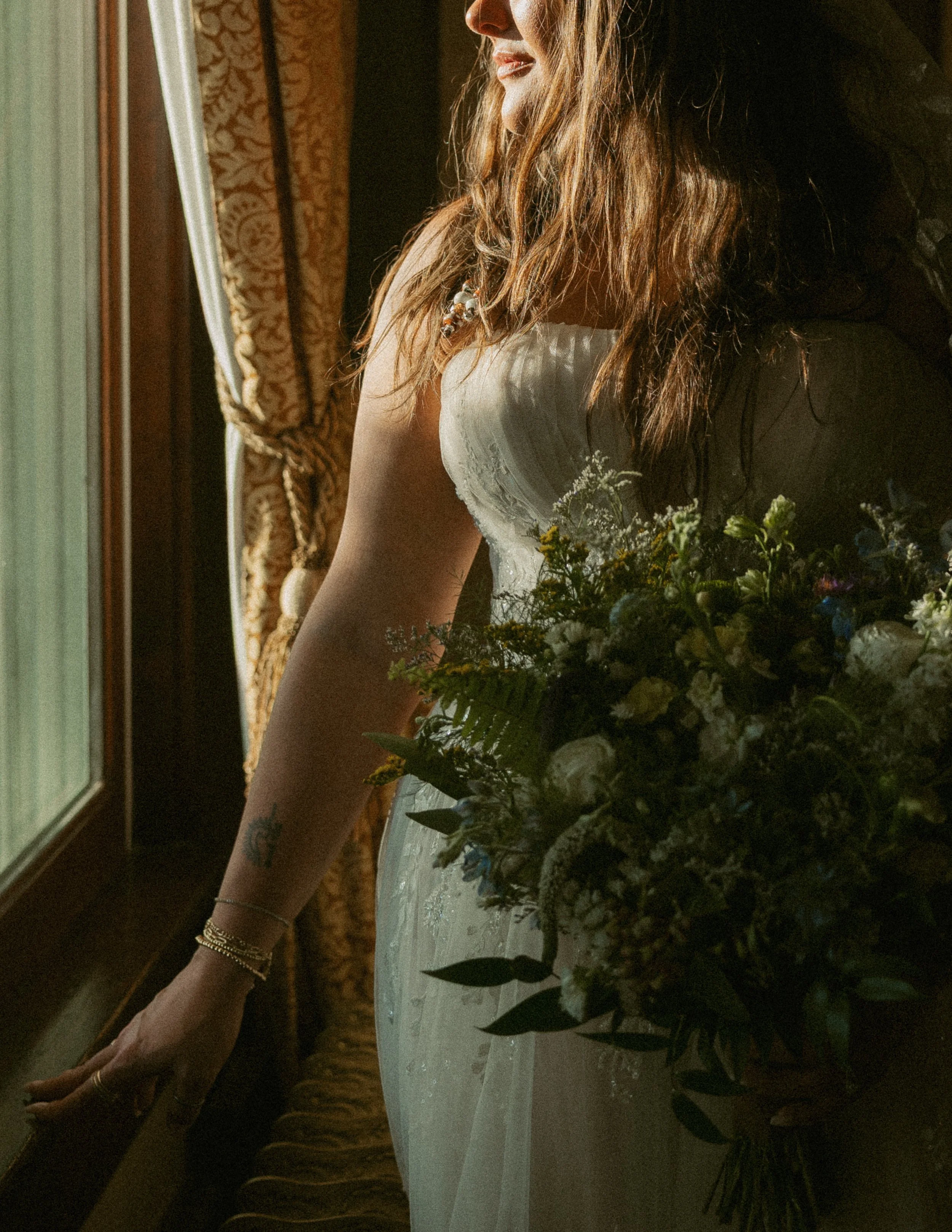 A woman in a white dress with floral details, wearing gold jewelry, is sitting at a table near a window with curtains. She is looking slightly to her left with sunlight illuminating her face and hair, and a bouquet of flowers is on the table.