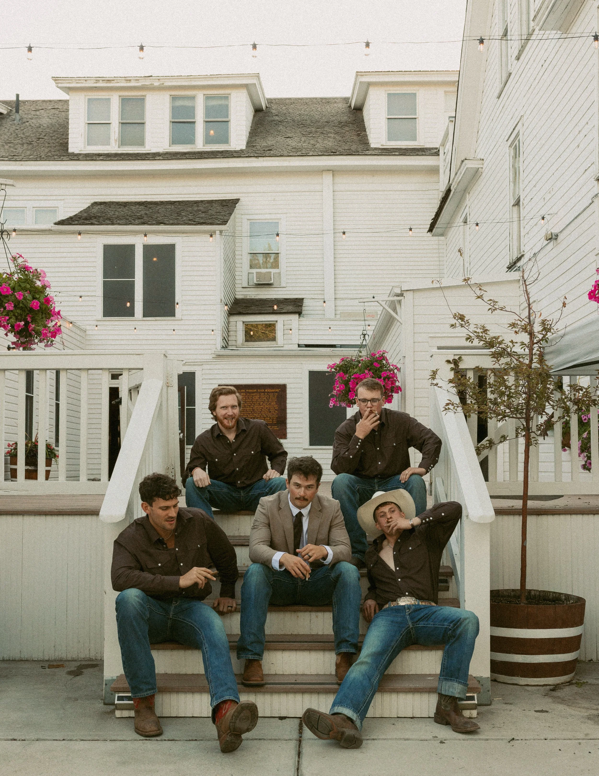 Group of people sitting on the steps of a white house, some are smiling and others are smoking, with string lights overhead and pink flowers nearby.