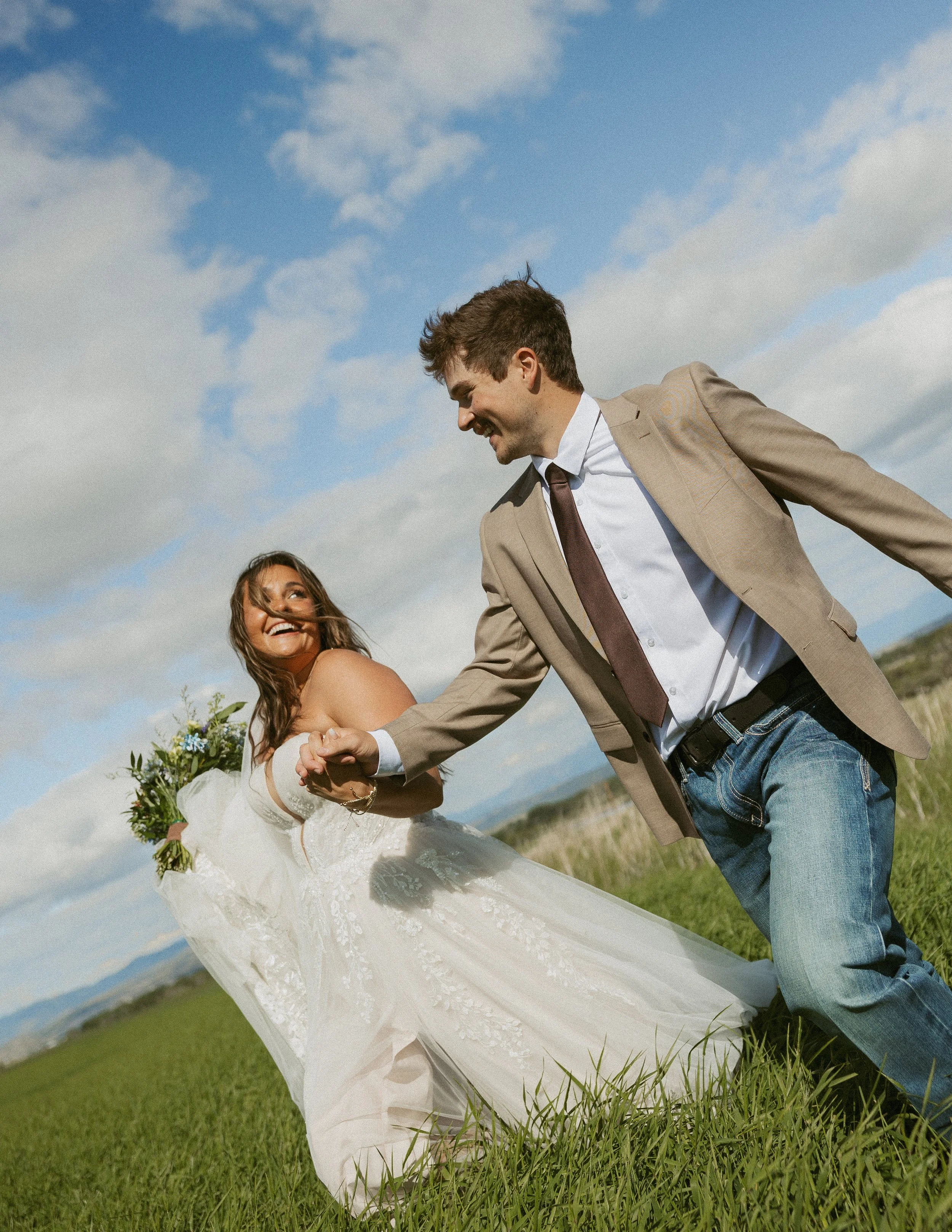 A bride and groom holding hands, smiling, outdoors on a grassy field with a blue sky and clouds.