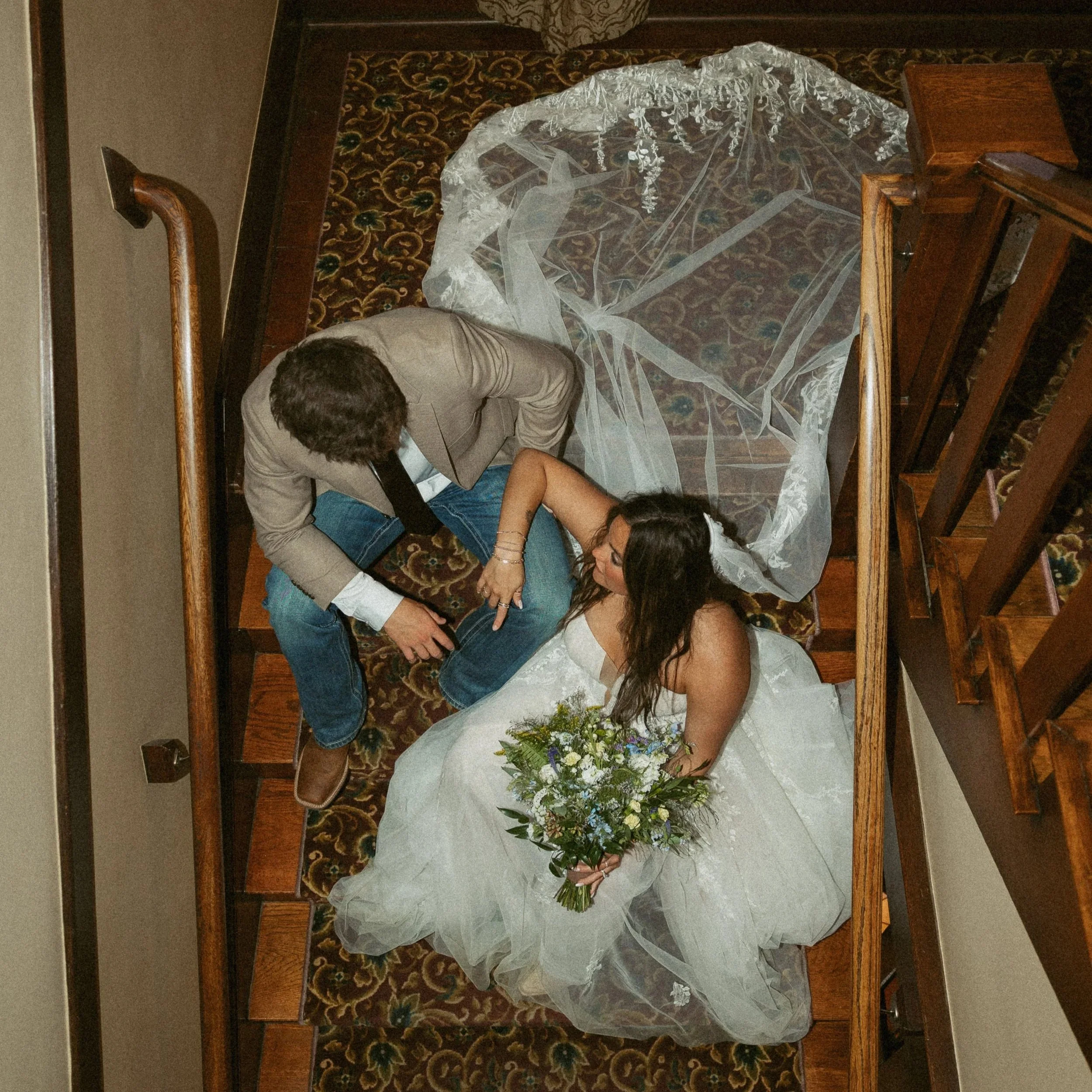 A wedding scene with a bride and groom lying on the floor, covered with a white bridal veil, surrounded by flowers, with guests nearby in a decorated venue.