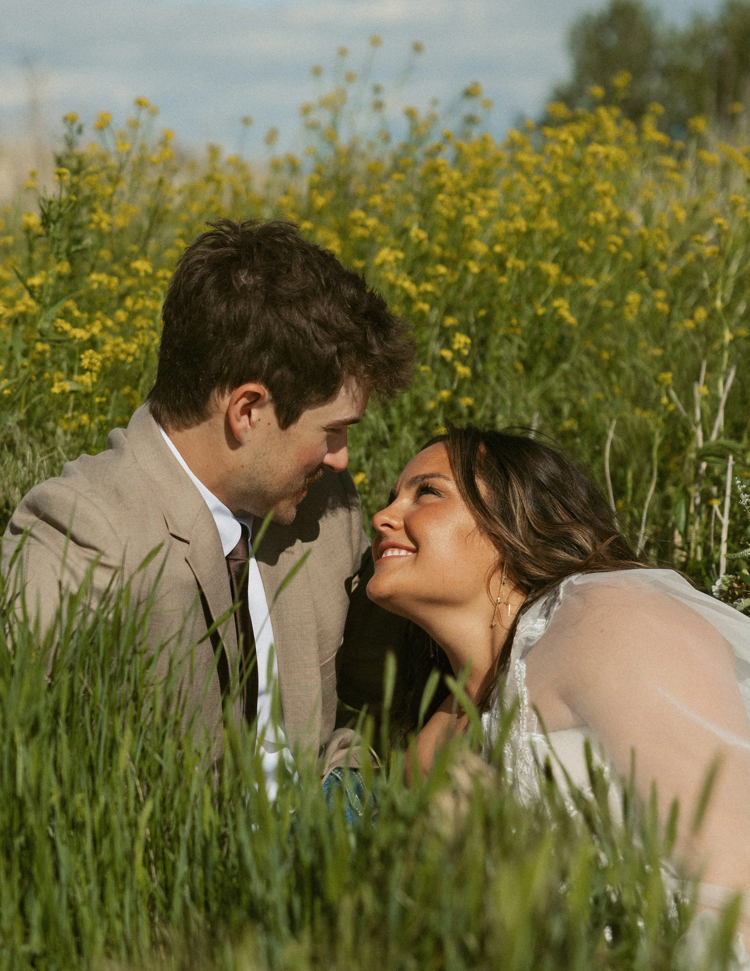 A man and a woman in wedding attire lying in a grassy field with yellow flowers, facing each other closely, with a background of green bushes and trees.