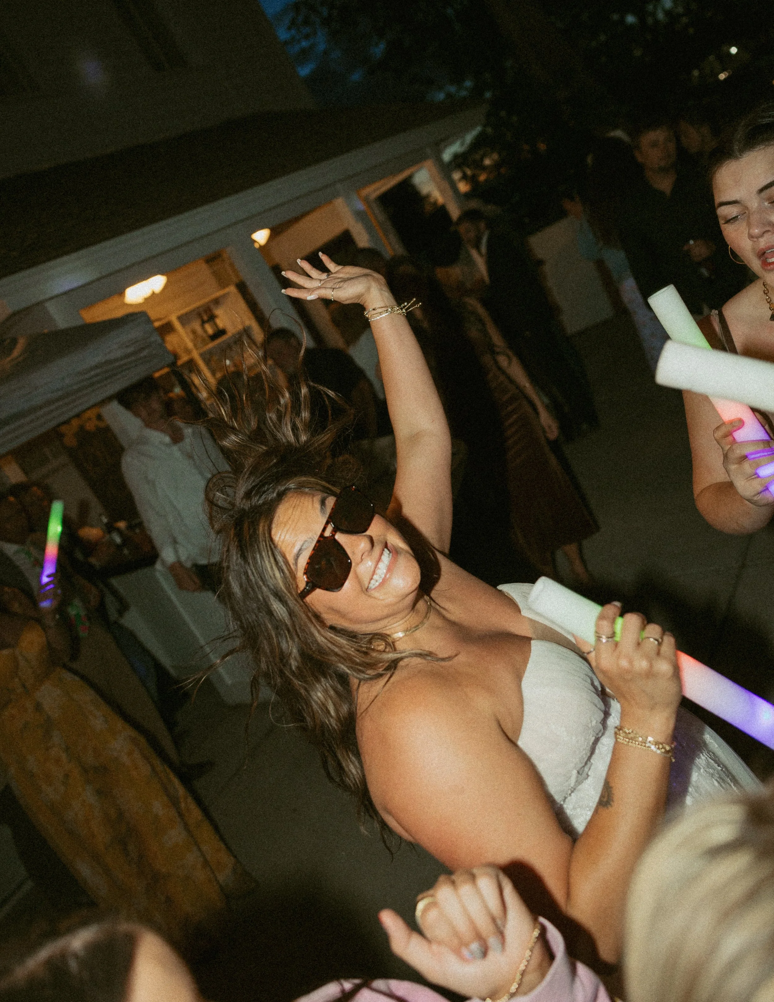 A woman dancing at a party, wearing sunglasses, a white dress, and gold jewelry, smiling with her arm raised. The background shows other guests, some talking and some dancing, in a dimly lit room.