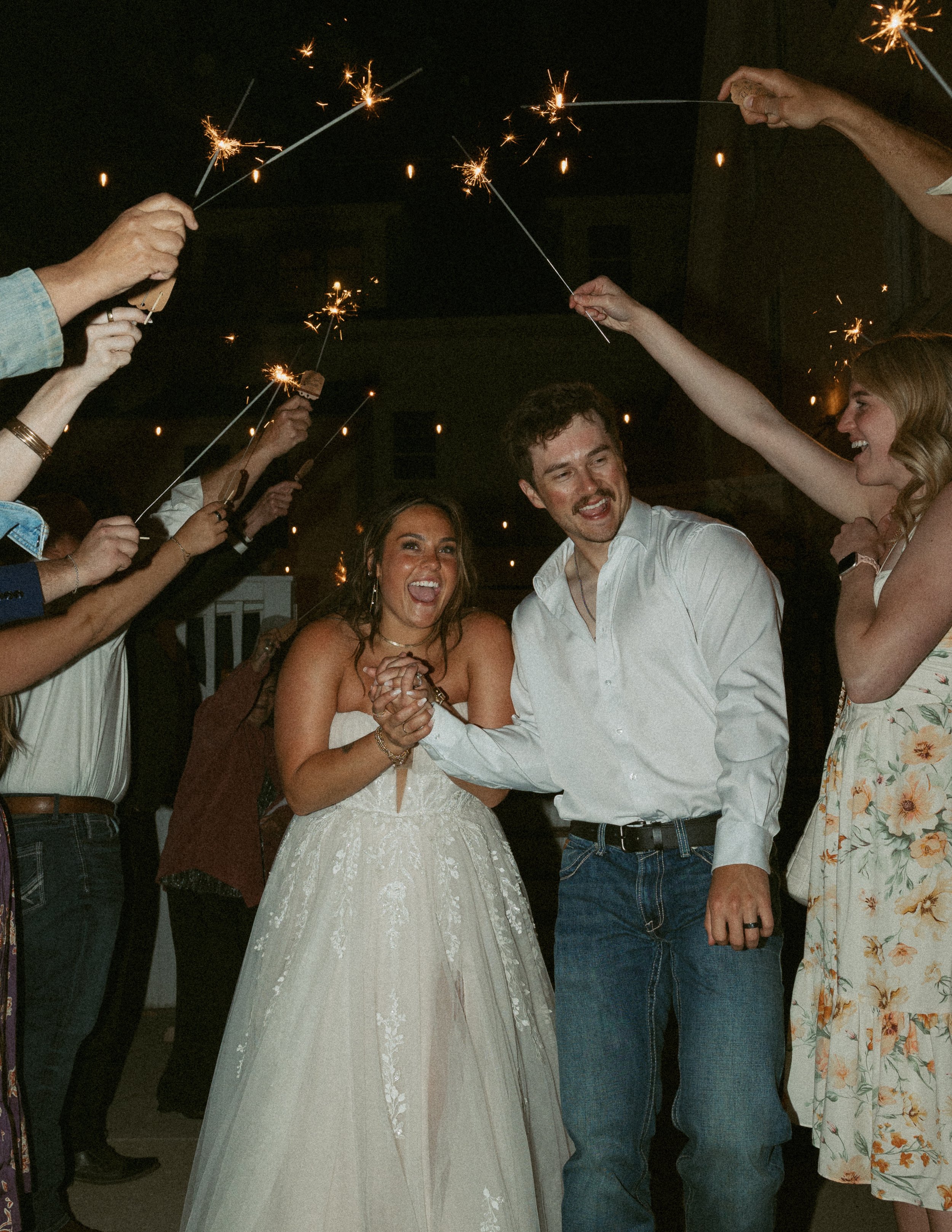 People celebrating with sparklers at night, two central individuals are dancing, a bride and groom, smiling and holding hands, surrounded by friends.