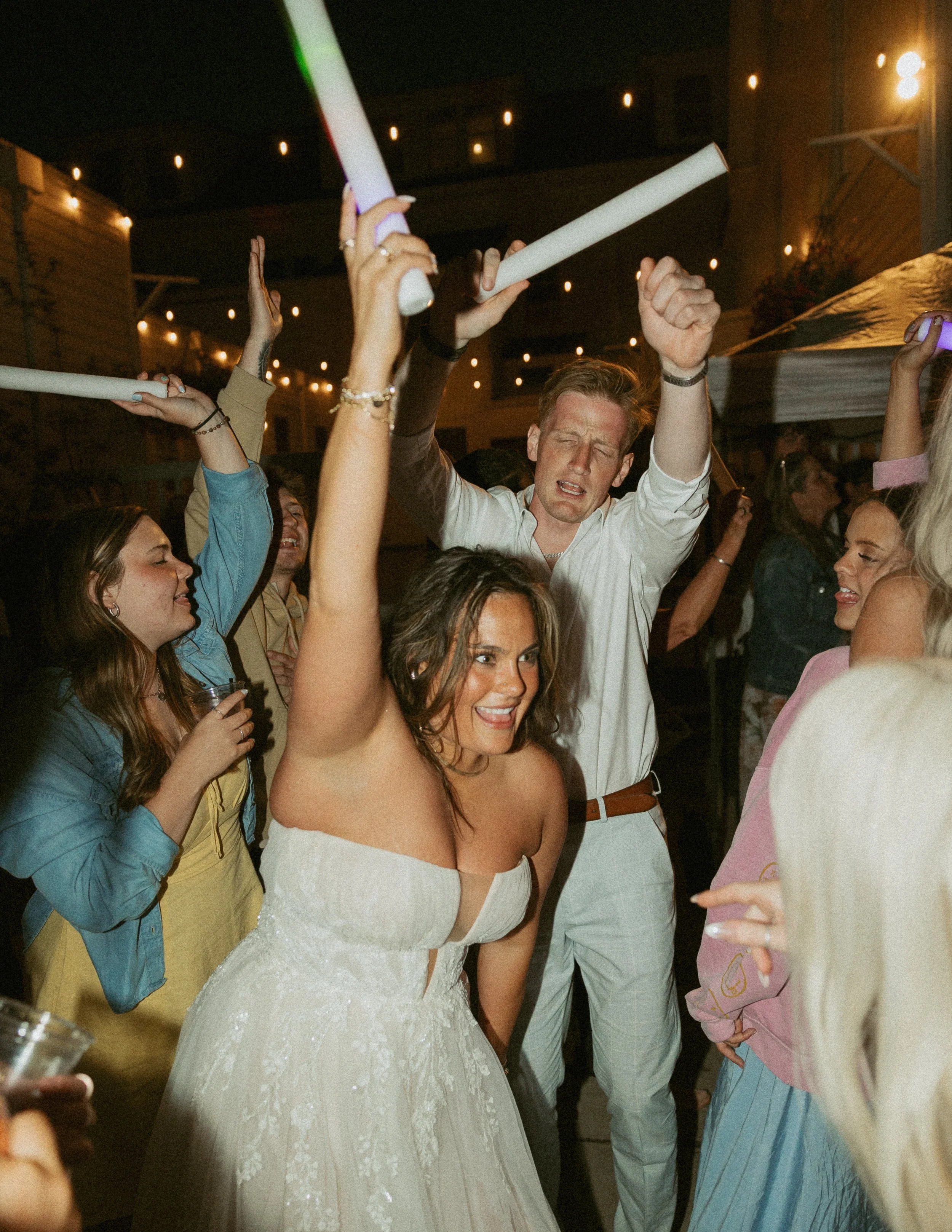 People dancing at a party, including a woman in a white lace dress and a man with closed eyes holding a glow stick.