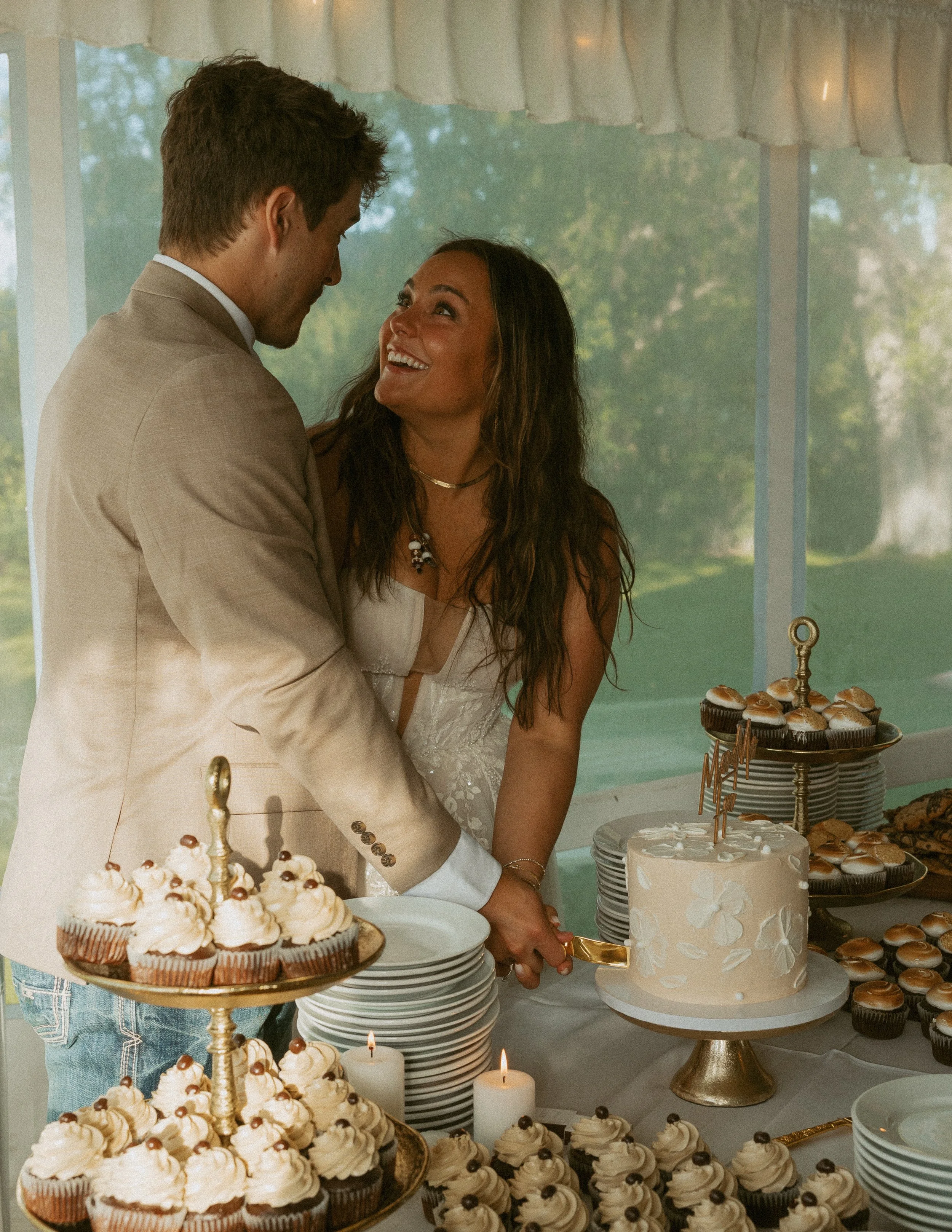 A newlywed couple at a wedding reception, standing behind a dessert table with cupcakes and a white wedding cake. The bride is smiling up at the groom. The table has lit candles and decorative desserts.