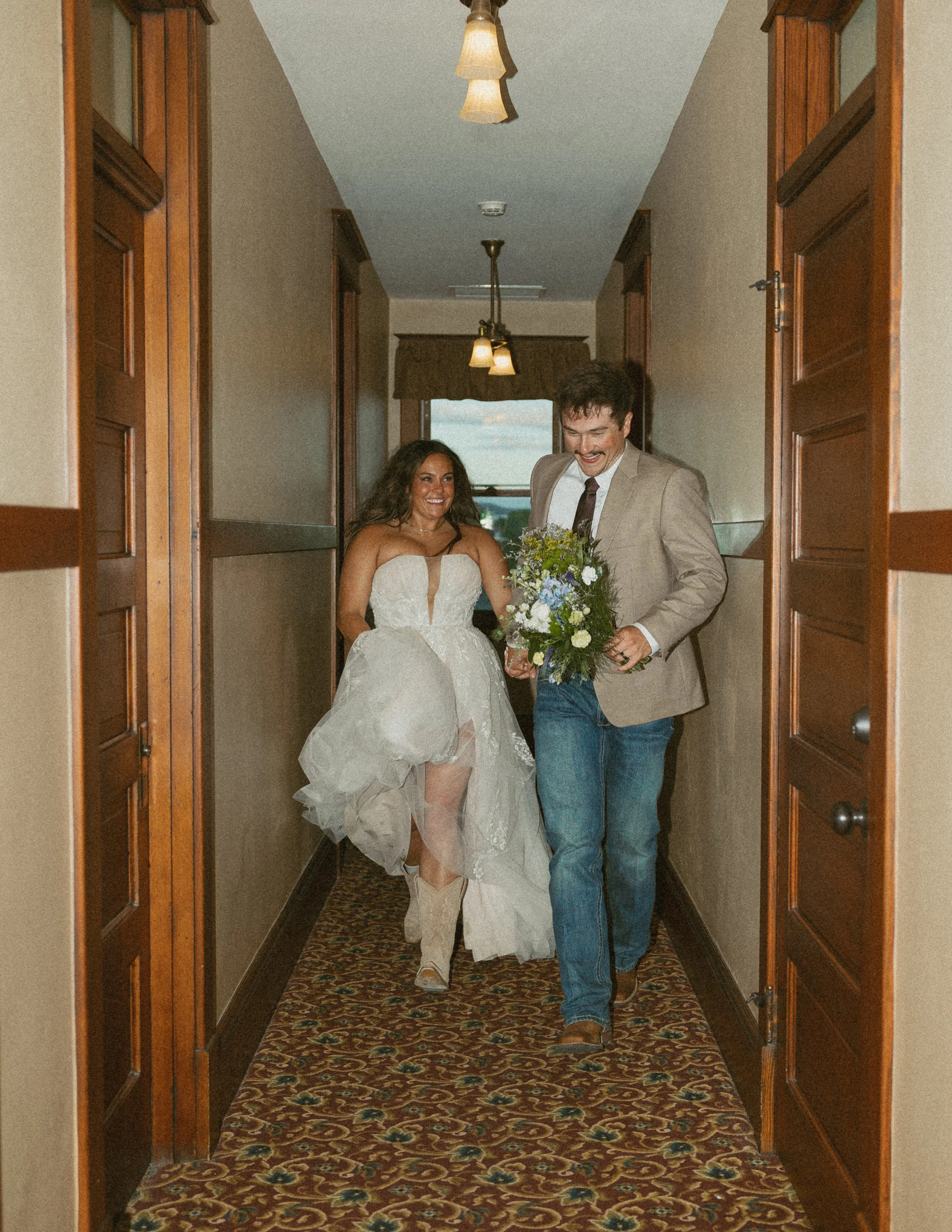 A bride in a white strapless wedding gown is holding hands with a man in a beige blazer, light shirt, dark tie, and jeans as they walk down a hallway of a hotel.