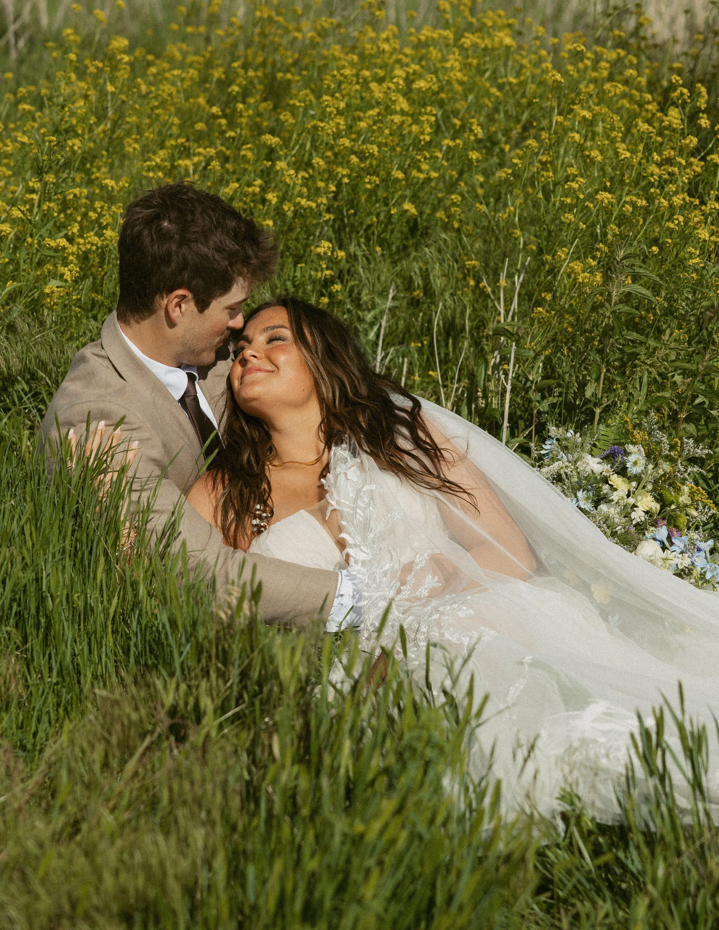 A couple laying in a field of tall grass and yellow flowers, smiling and looking at each other.