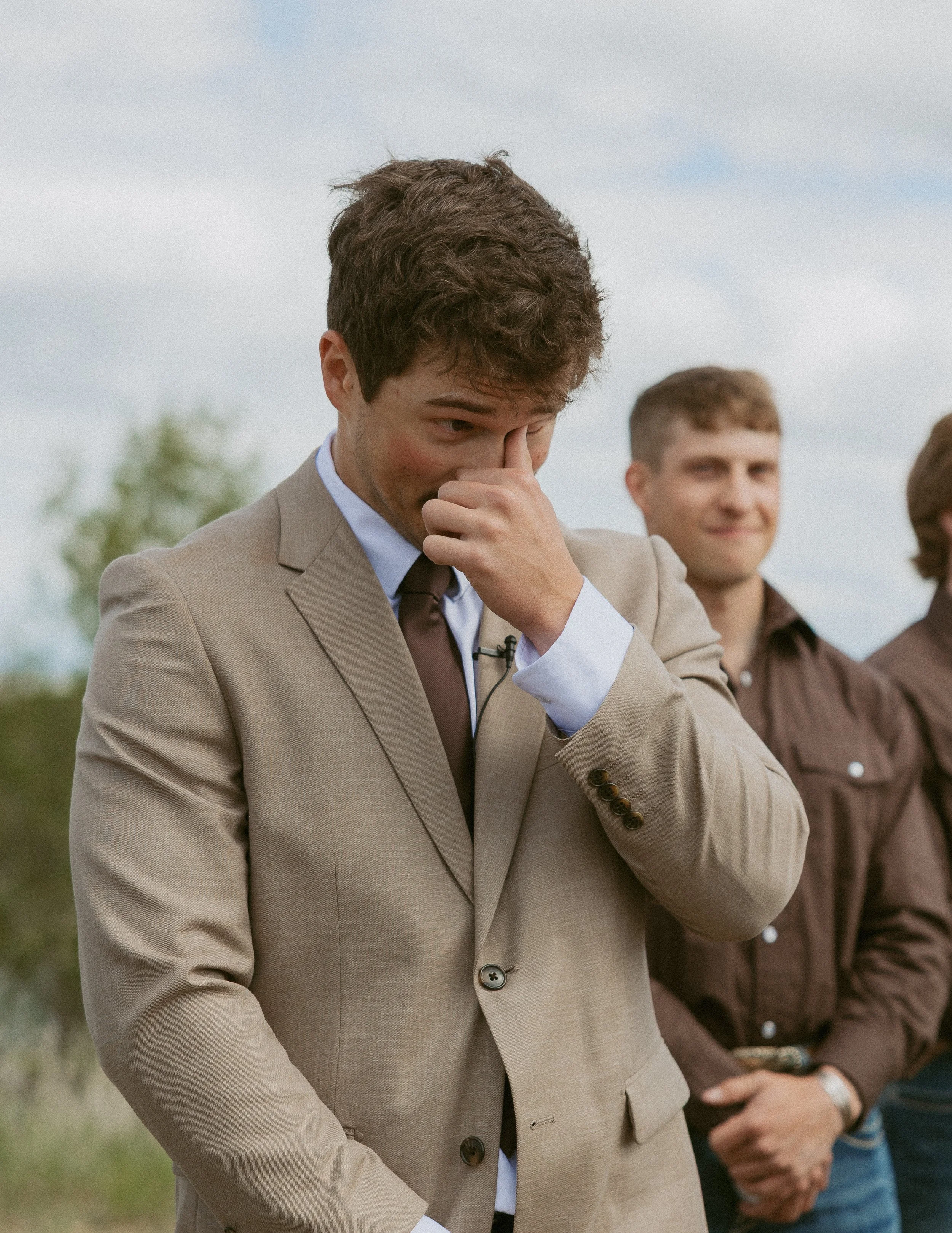 A young man in a beige suit covering his nose with his hand, standing outdoors against a cloudy sky with two other men in casual clothing in the background.