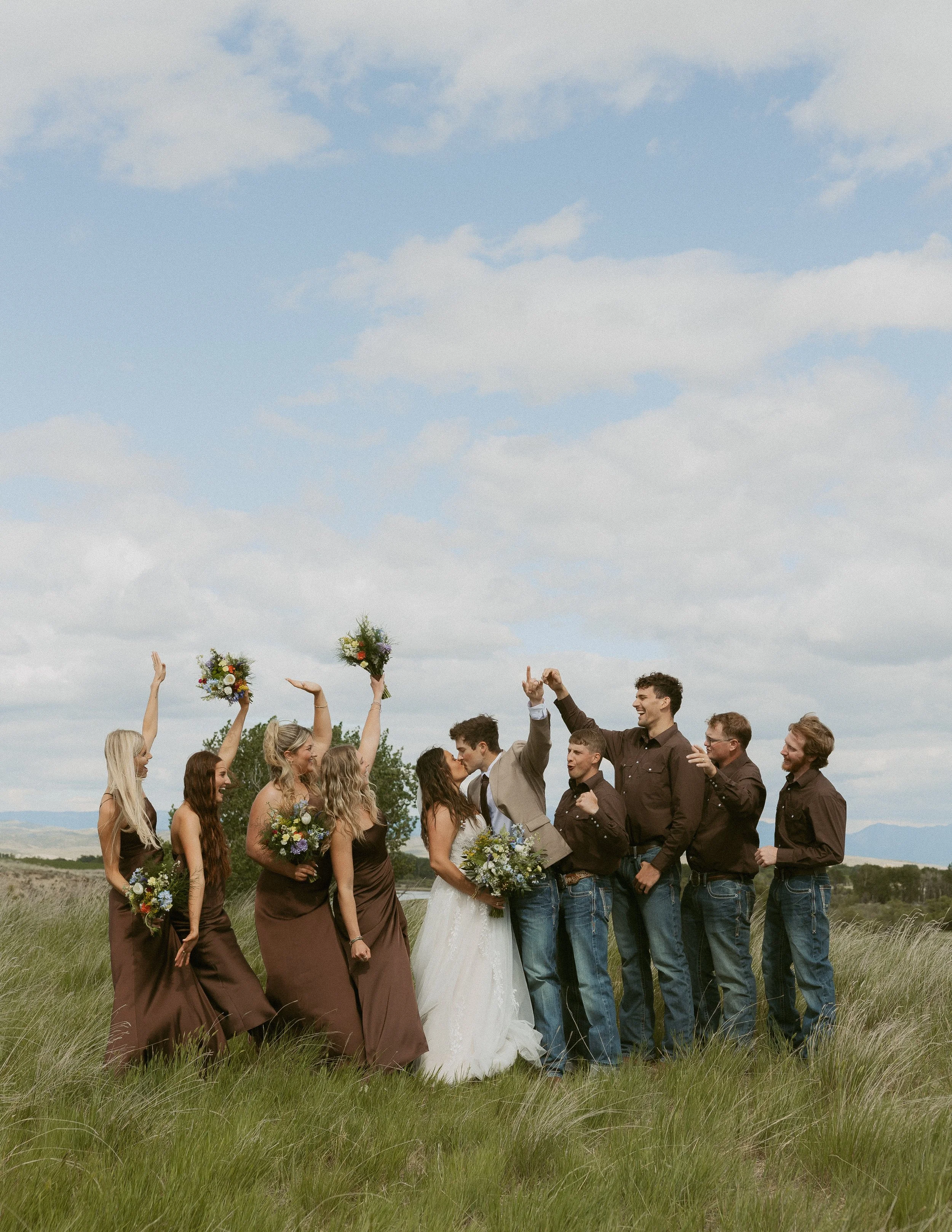 A group of people celebrating a wedding outdoors, with some holding bouquets and others raising their hands in the air, under a partly cloudy sky over a grassy field.