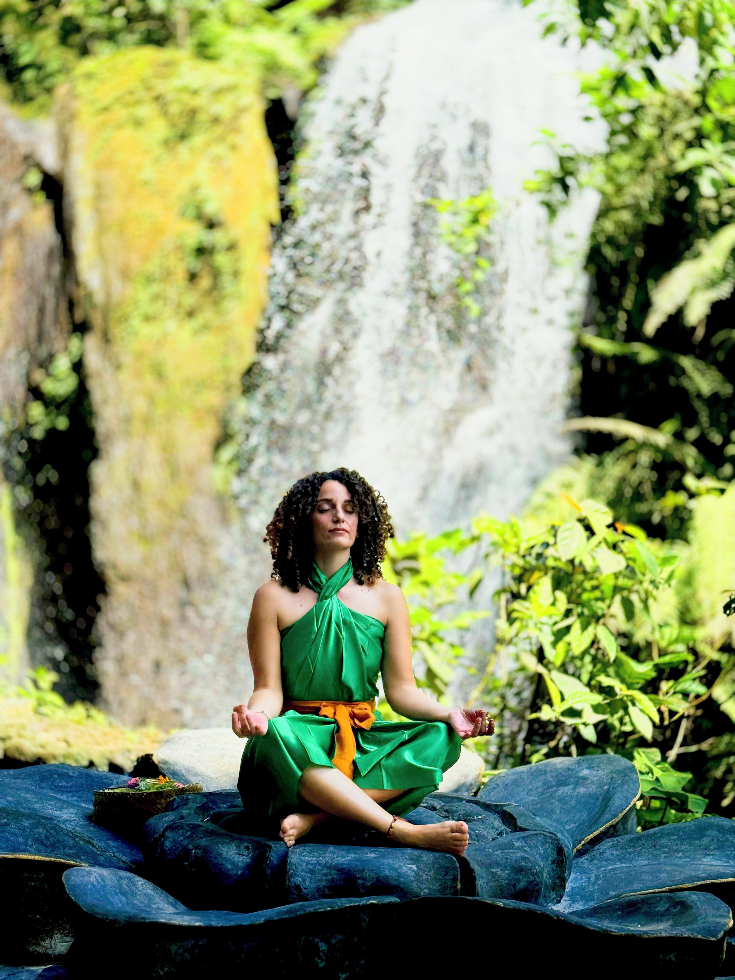 A woman in a green dress practicing yoga outdoors on a large flat rock, with a waterfall and lush greenery in the background.