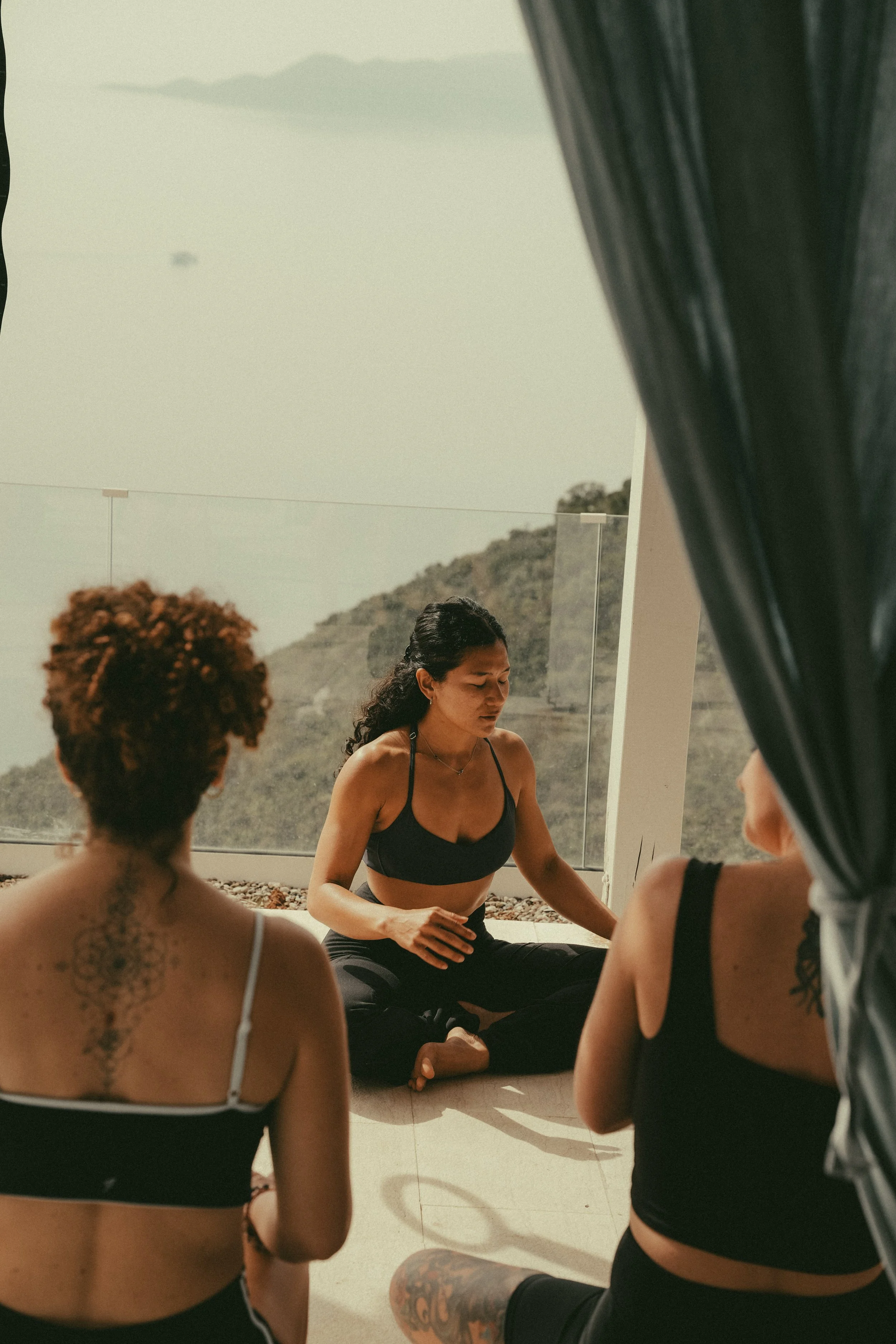 Women participating in a yoga or meditation session, sitting on the floor inside a well-lit room with large windows, overlooking hills and trees outside.