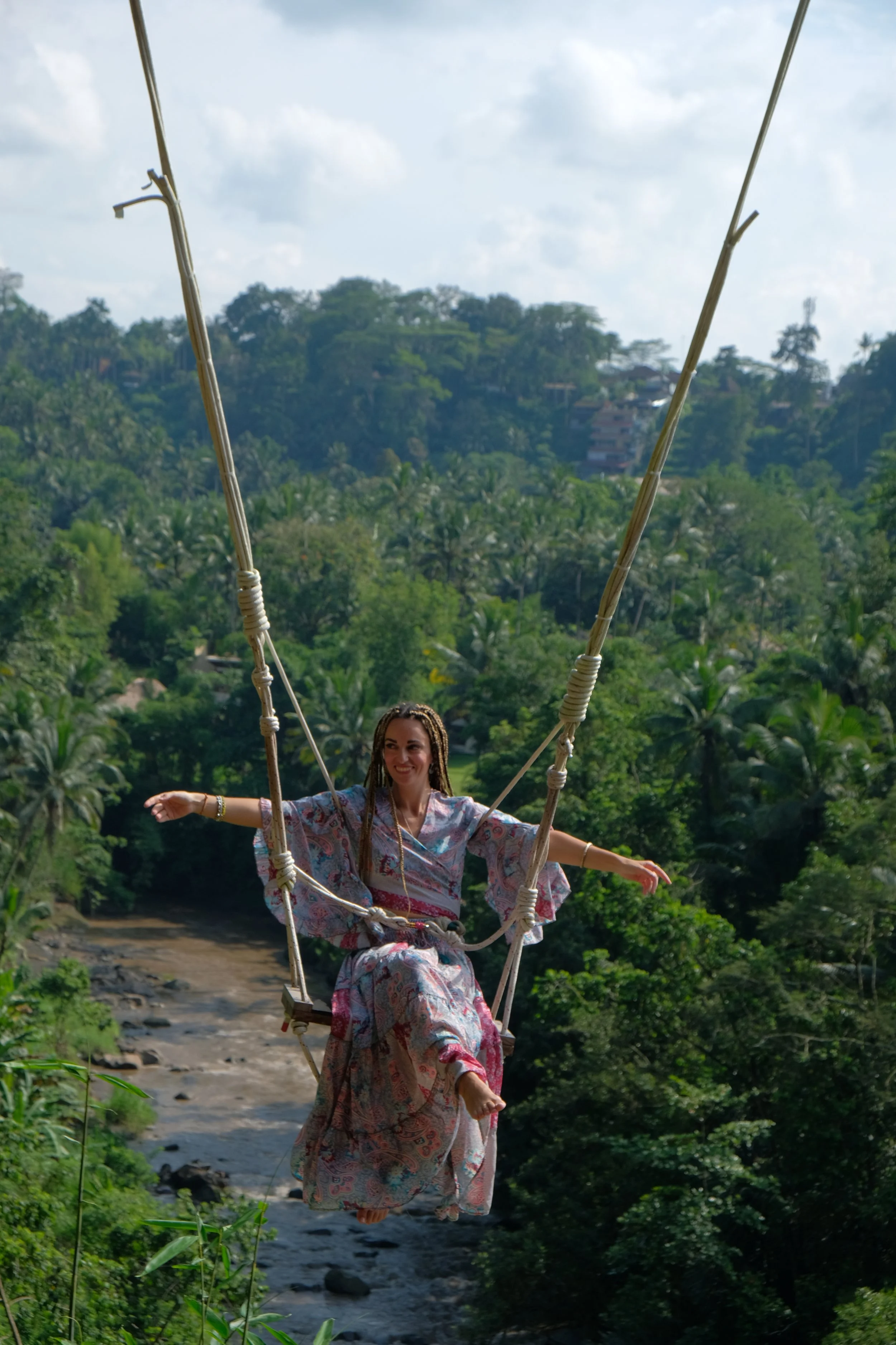 A woman in a floral kimono-style dress with dreadlocks is smiling and sitting on a swing over a river, surrounded by lush green trees and hills in the background.