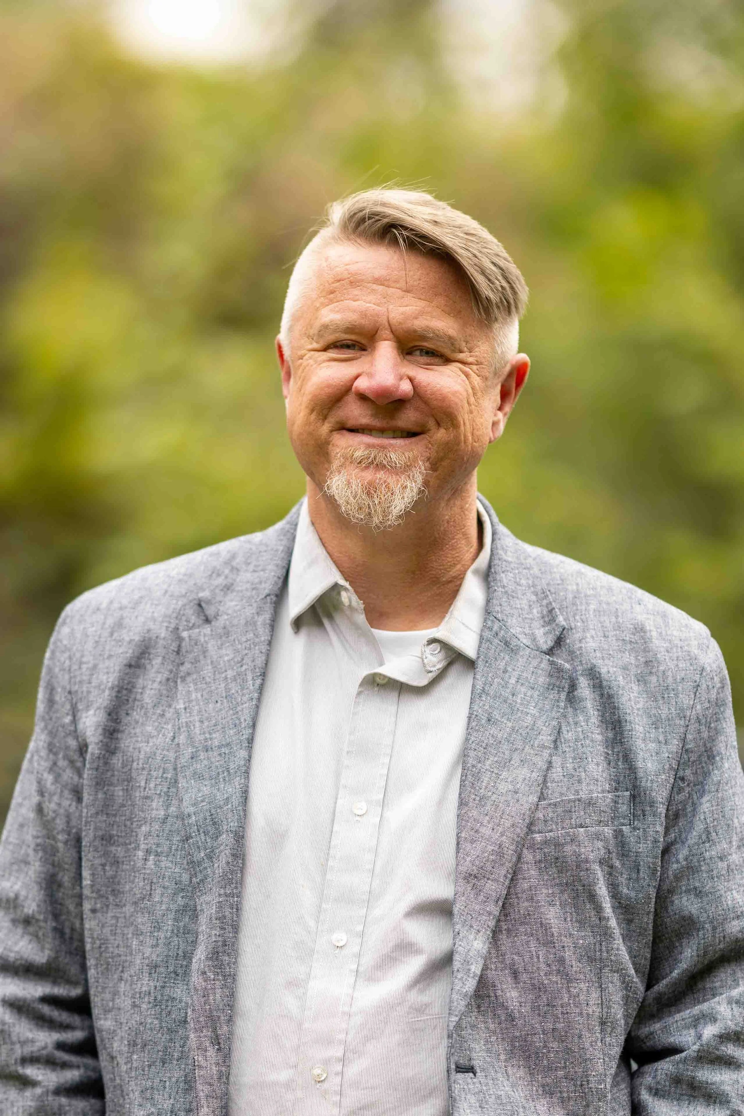 Outdoor portrait of a middle-aged man with short blonde hair, a goatee, and a slight smile, wearing a gray blazer over a white button-up shirt, with a green, blurred natural background.