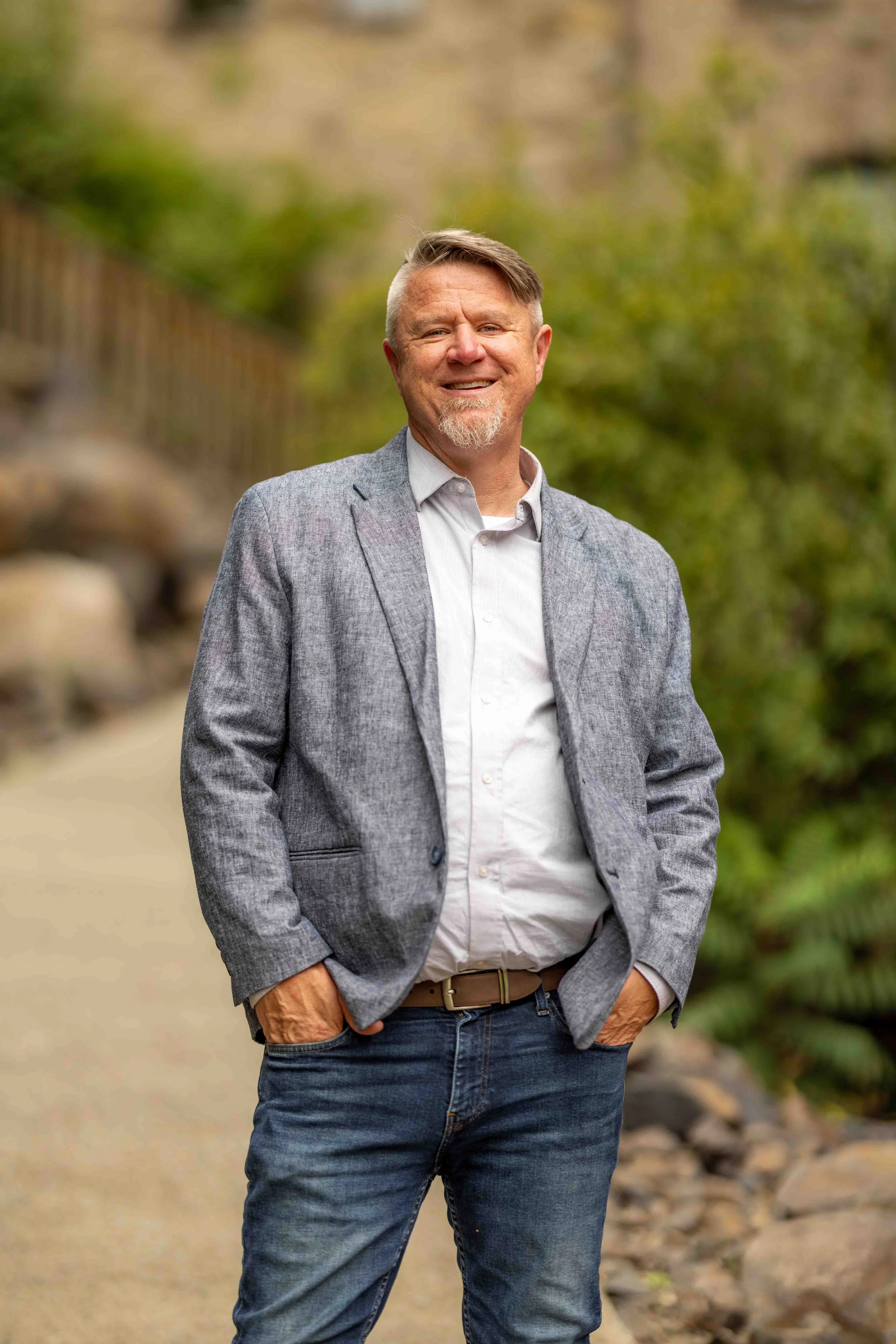A smiling middle-aged man with short, light brown hair and a beard, wearing a light gray blazer, white button-up shirt, and blue jeans, standing outdoors on a rocky trail with a blurred background of green trees and rocks.