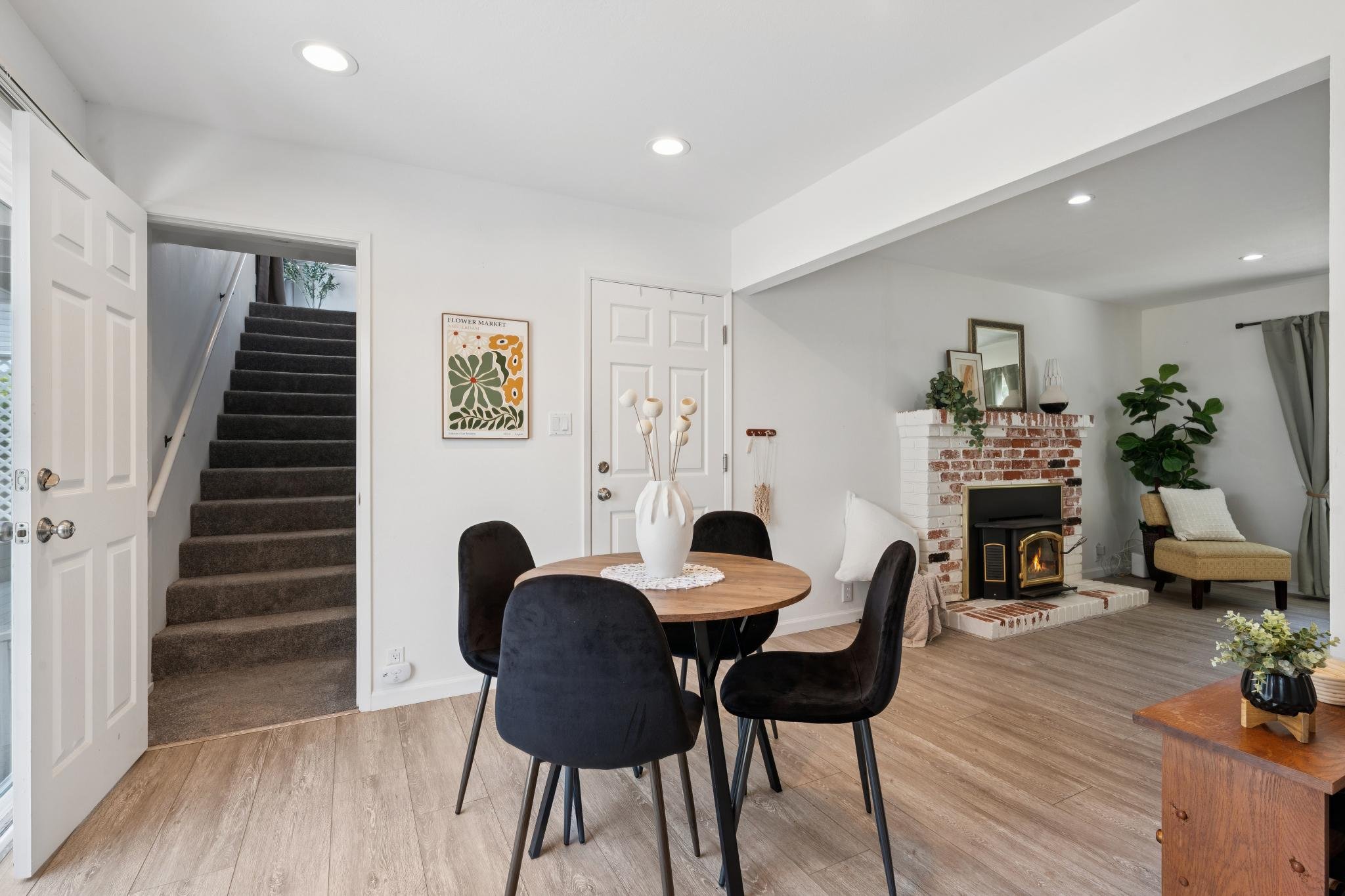 Bright living room with white walls, a fireplace with a brick surround, a small plant on the mantel, a mirror, and a chair, with a dining table and black chairs in the foreground. Stairs with dark carpet leading upstairs on the left side.