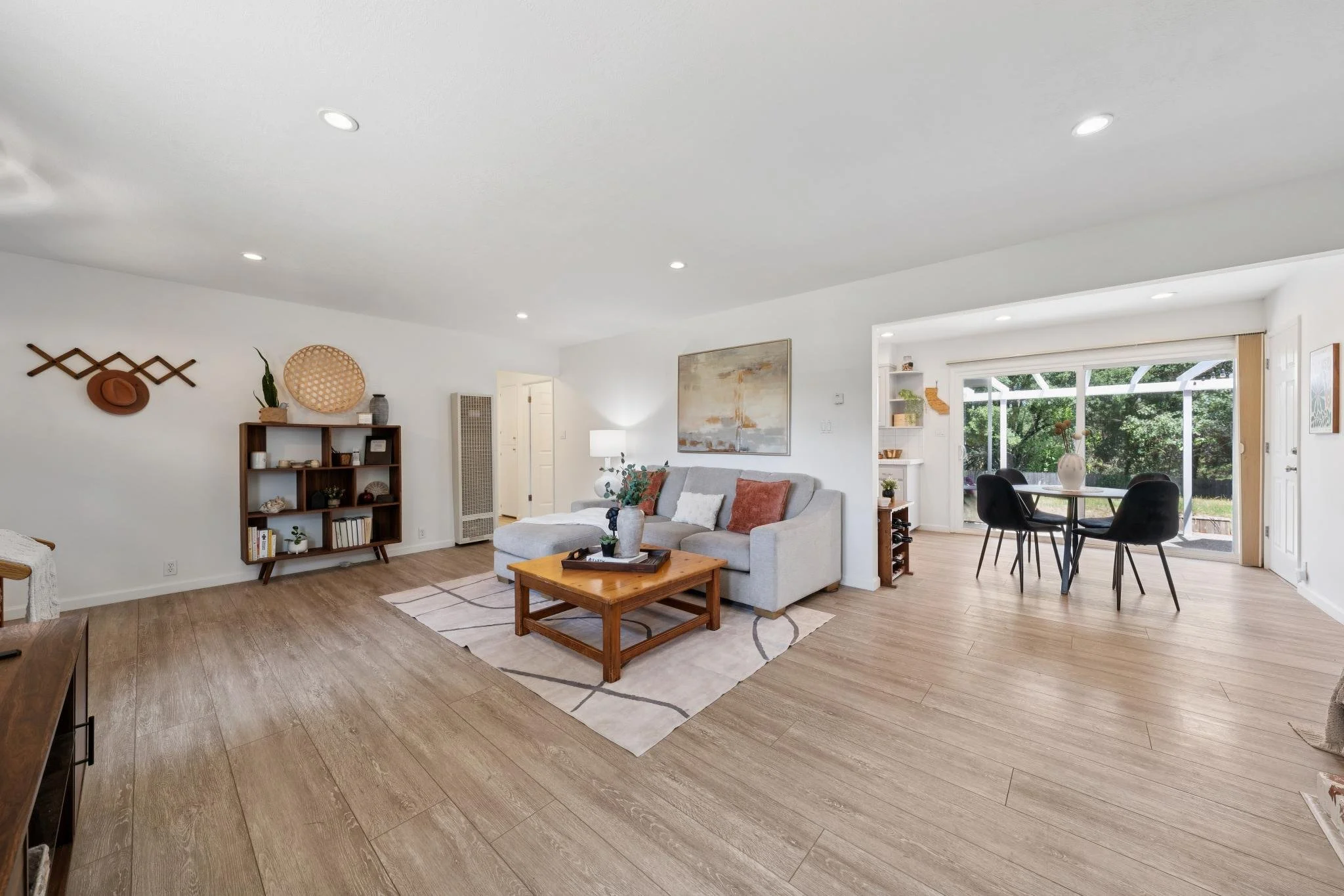 Modern living room with white walls, wooden flooring, a beige sofa with orange and white pillows, a wooden coffee table, a tall lamp, a wall art, and built-in shelves near sliding glass doors showing an outdoor patio with trees.