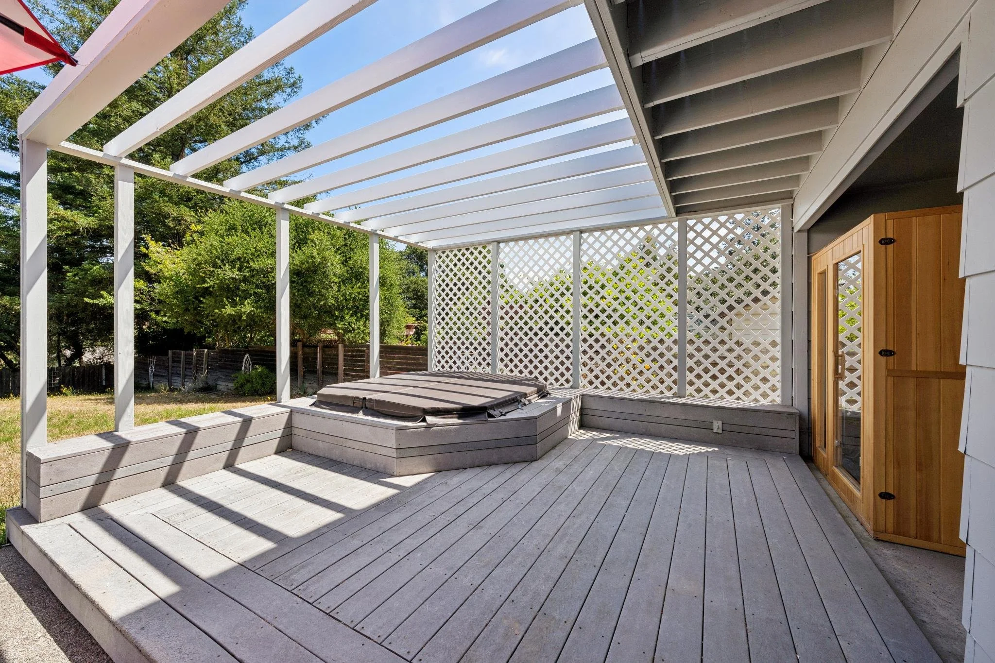 Back patio with white wooden decking, lattice fencing, and privacy screen, featuring a hot tub and a wooden storage shed in a backyard with trees.